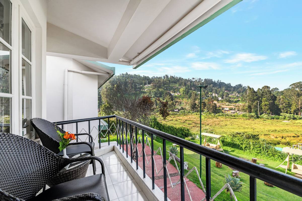 Un balcon agrémenté de fauteuils et d’une table en osier, d’où l’on peut admirer un paysage verdoyant et luxuriant, avec des arbres et un village situé au loin.