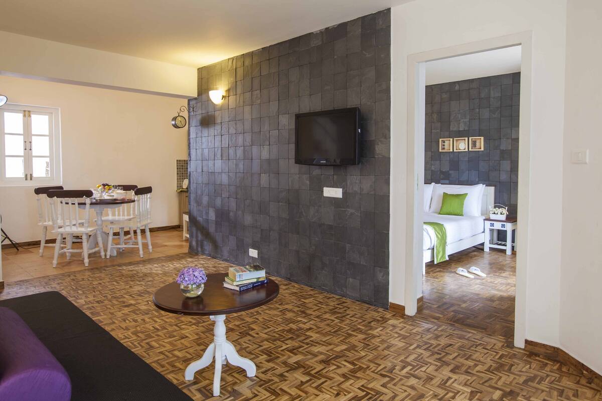 Un salon doté d’un parquet en bois disposé en chevrons, d’un canapé blanc, d’une petite table ronde surmontée d’un vase, d’un coin repas avec une table et des chaises blanches, et d’un mur recouvert de carreaux foncés sur lequel est fixée une télévision.