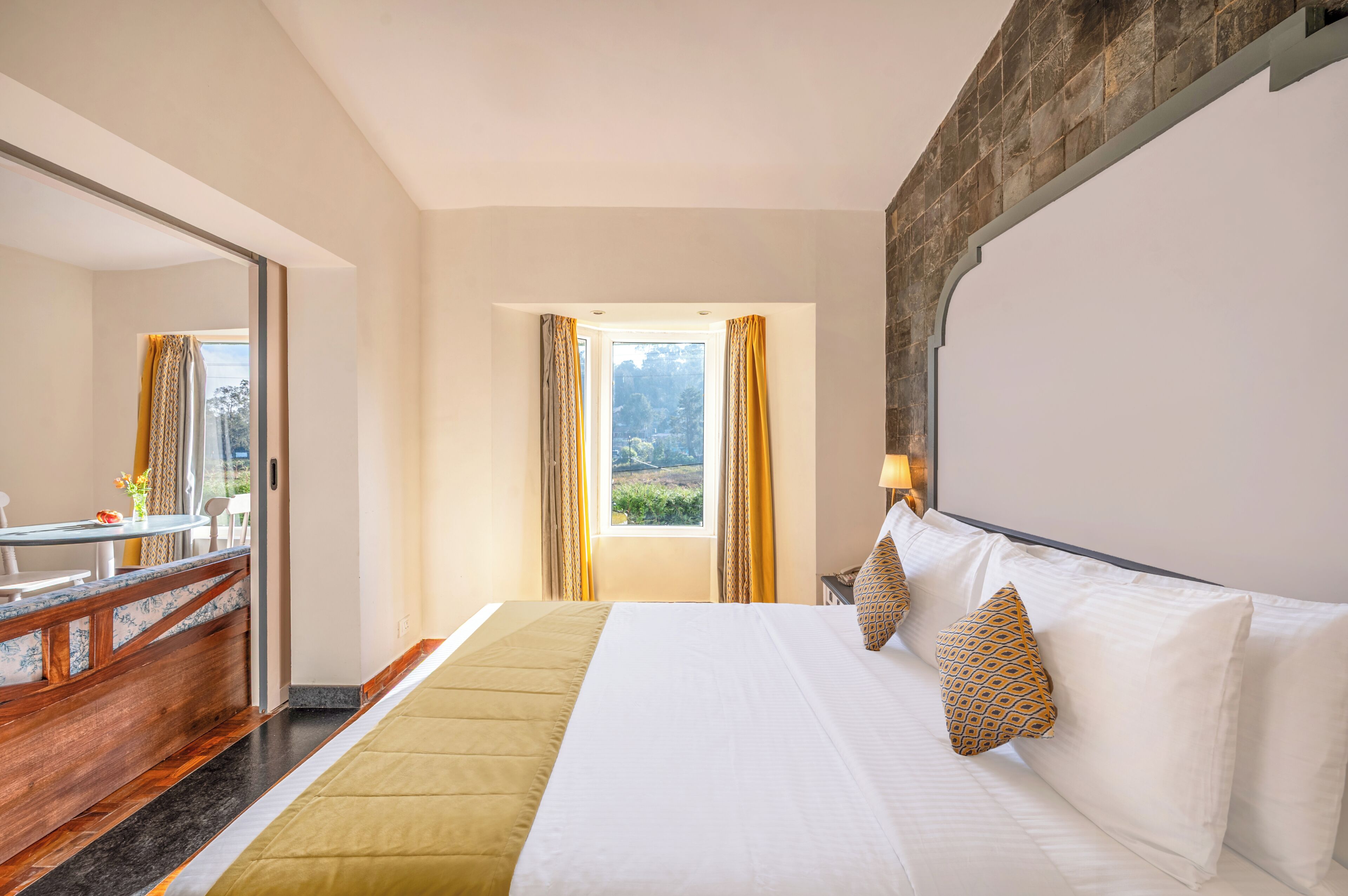 A hotel room with a bed, a desk, a chair, a window with curtains, and a view of greenery outside.