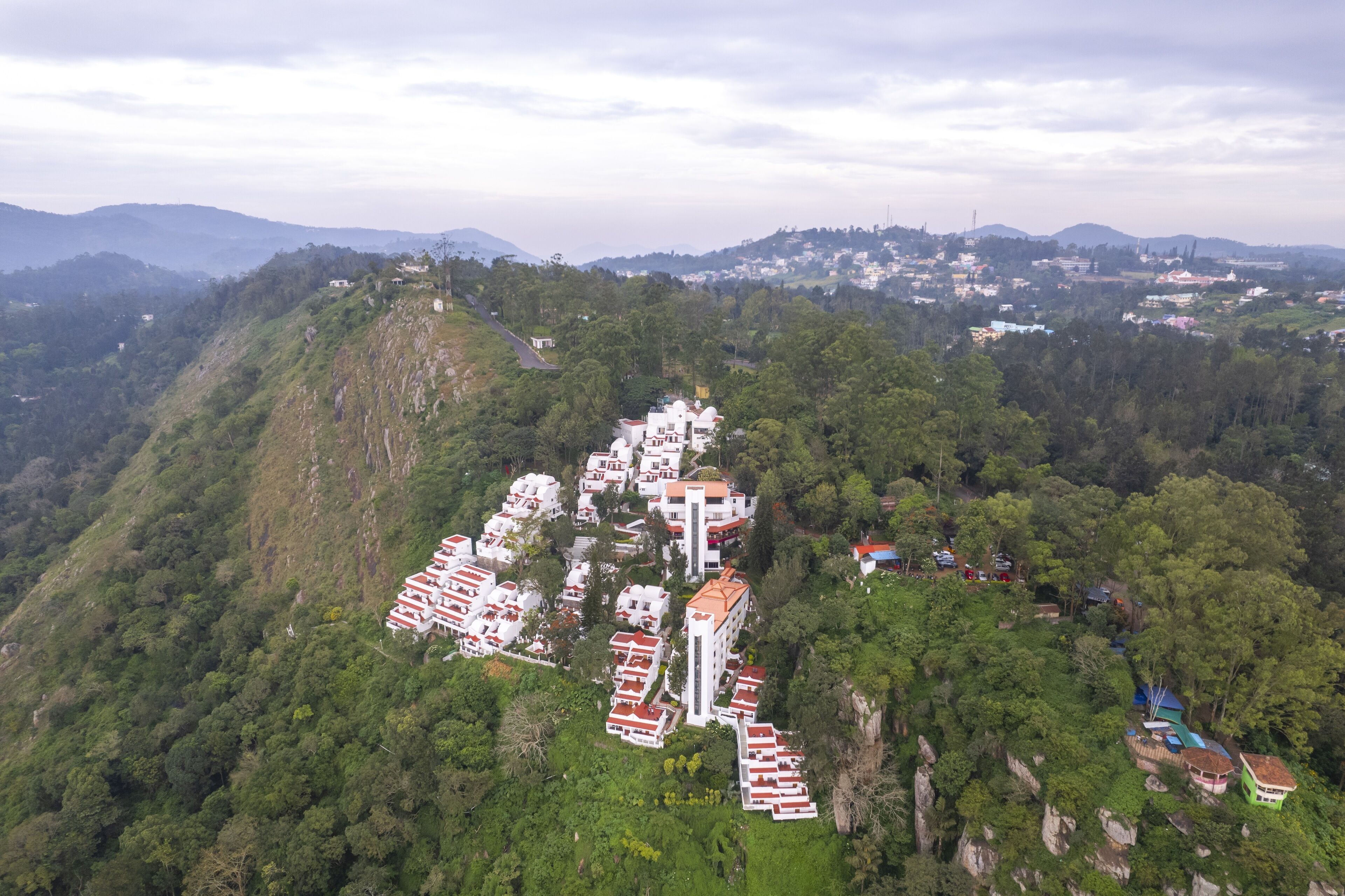 A hillside residential area with multiple buildings, some with red roofs, surrounded by dense greenery and hills in the background.