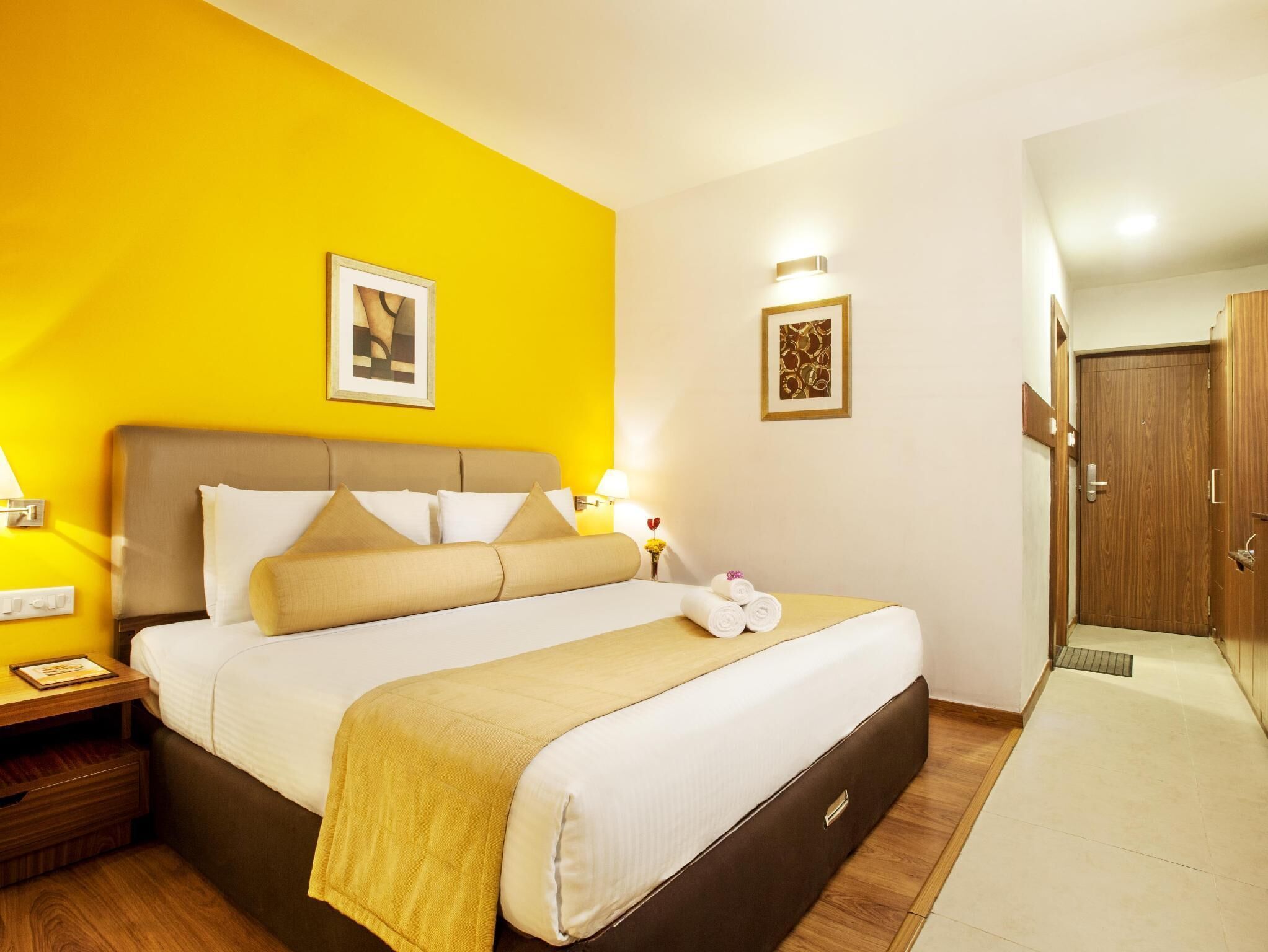 A hotel room with a bed, bedside tables, a nightstand, a wooden door, and a wall with two framed pictures.