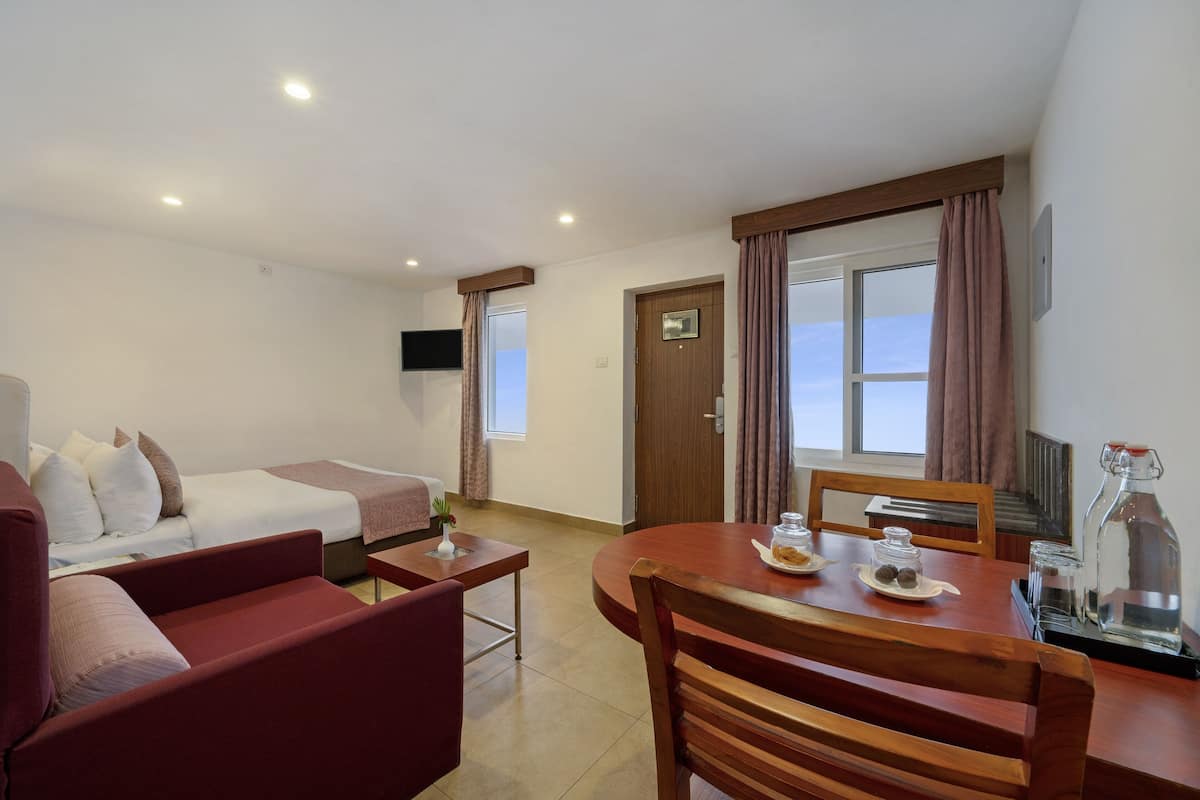 Une chambre d’hôtel comprenant un lit, un canapé, une table à manger avec des chaises, un téléviseur fixé au mur et une fenêtre avec des rideaux.