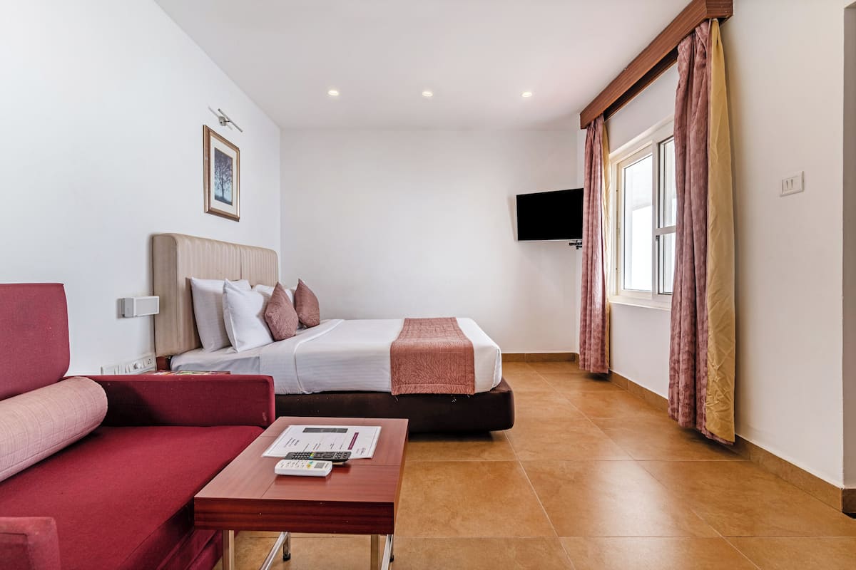 Une chambre d’hôtel avec un lit, un canapé rouge, une table en bois, un téléviseur fixé au mur et une fenêtre avec des rideaux.