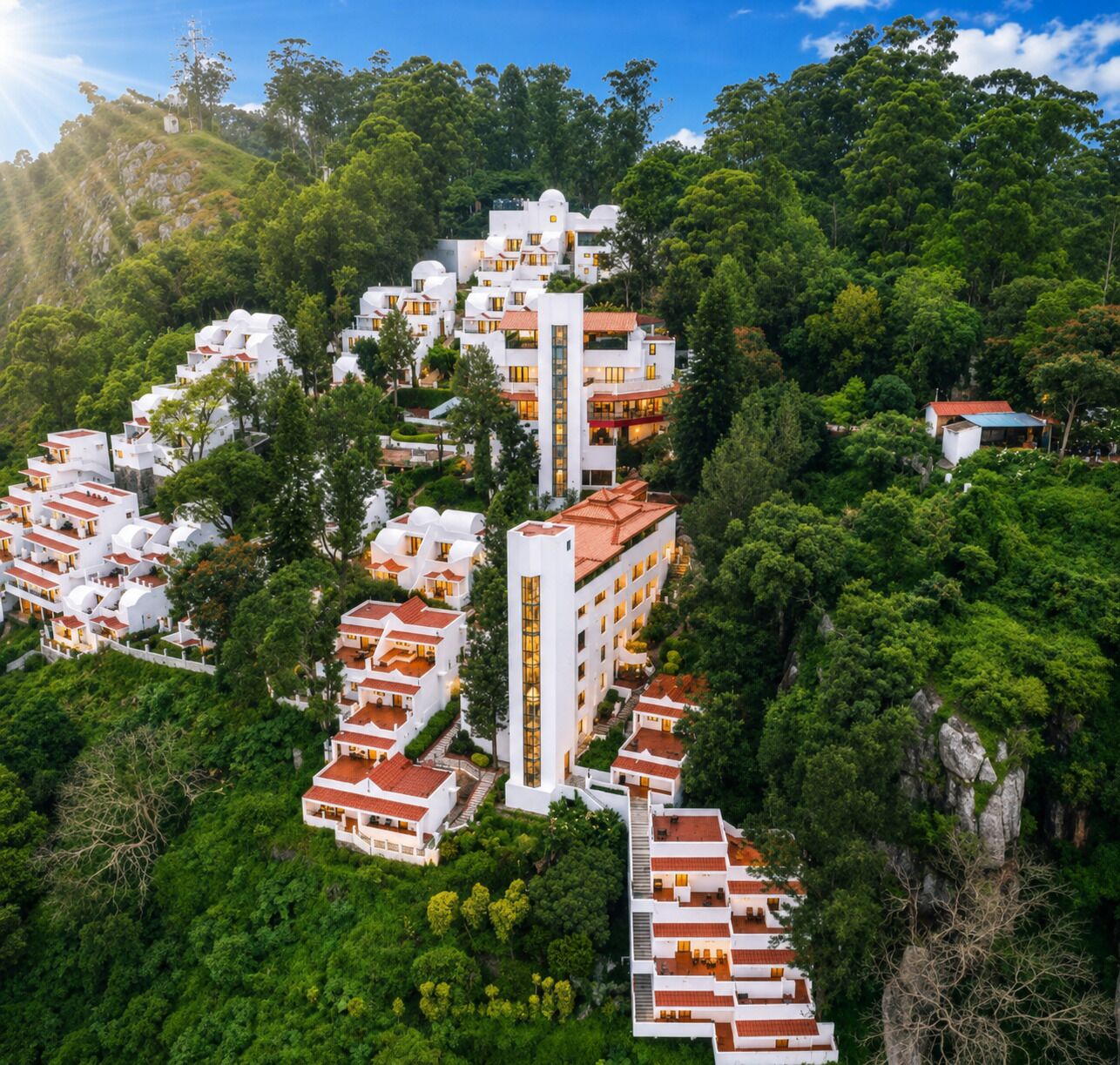 A hillside residential area with white buildings, some with red roofs, surrounded by dense green trees.