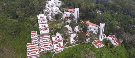 An aerial view of a residential area with white buildings and red roofs, surrounded by dense greenery and hills in the background.