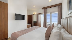 Une chambre d’hôtel avec un grand lit, un téléviseur fixé au mur, des meubles en bois et une vue sur le ciel par la fenêtre.