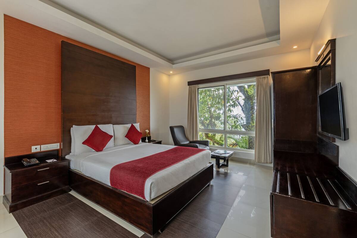 A modern hotel room with a large bed, a TV, a desk, and a window with a view of greenery.