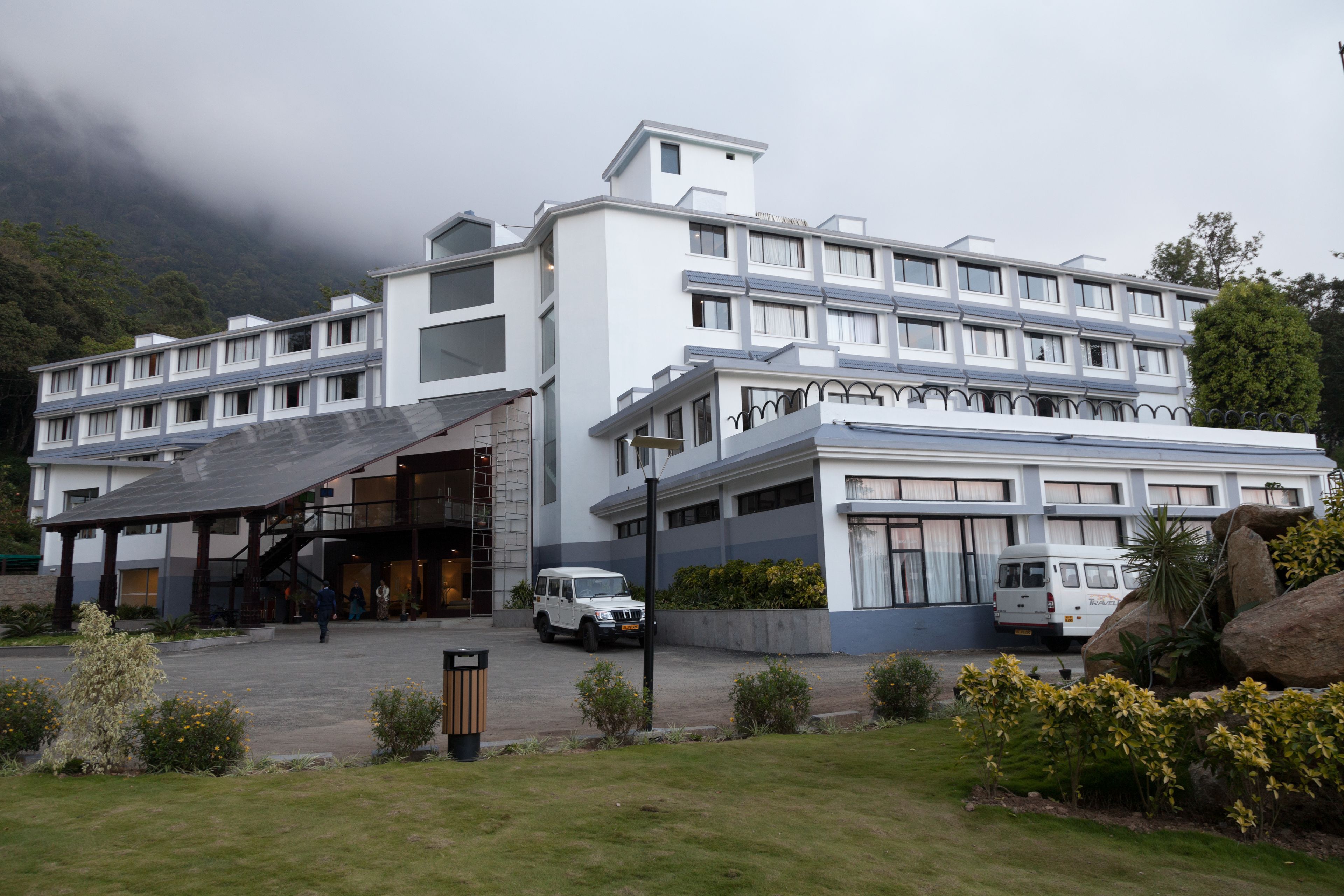 Un hôtel à plusieurs étages, doté d’un design moderne, avec une entrée couverte et un espace de stationnement où se trouvent des véhicules.