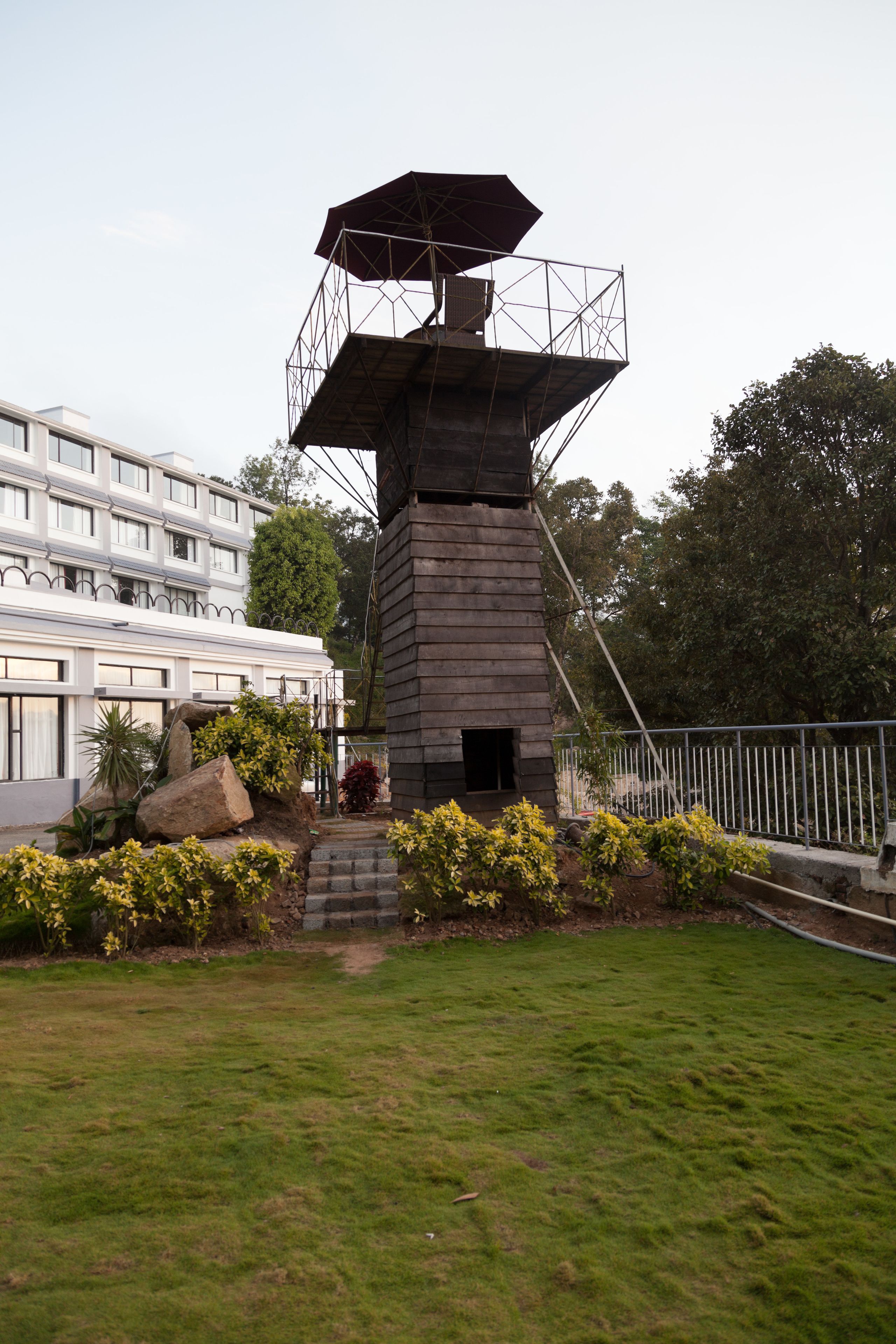 Una alta estructura de torre rústica con armazón metálico y una gran parte superior similar a una sombrilla, rodeada de un césped cuidado y un edificio con varias ventanas al fondo.