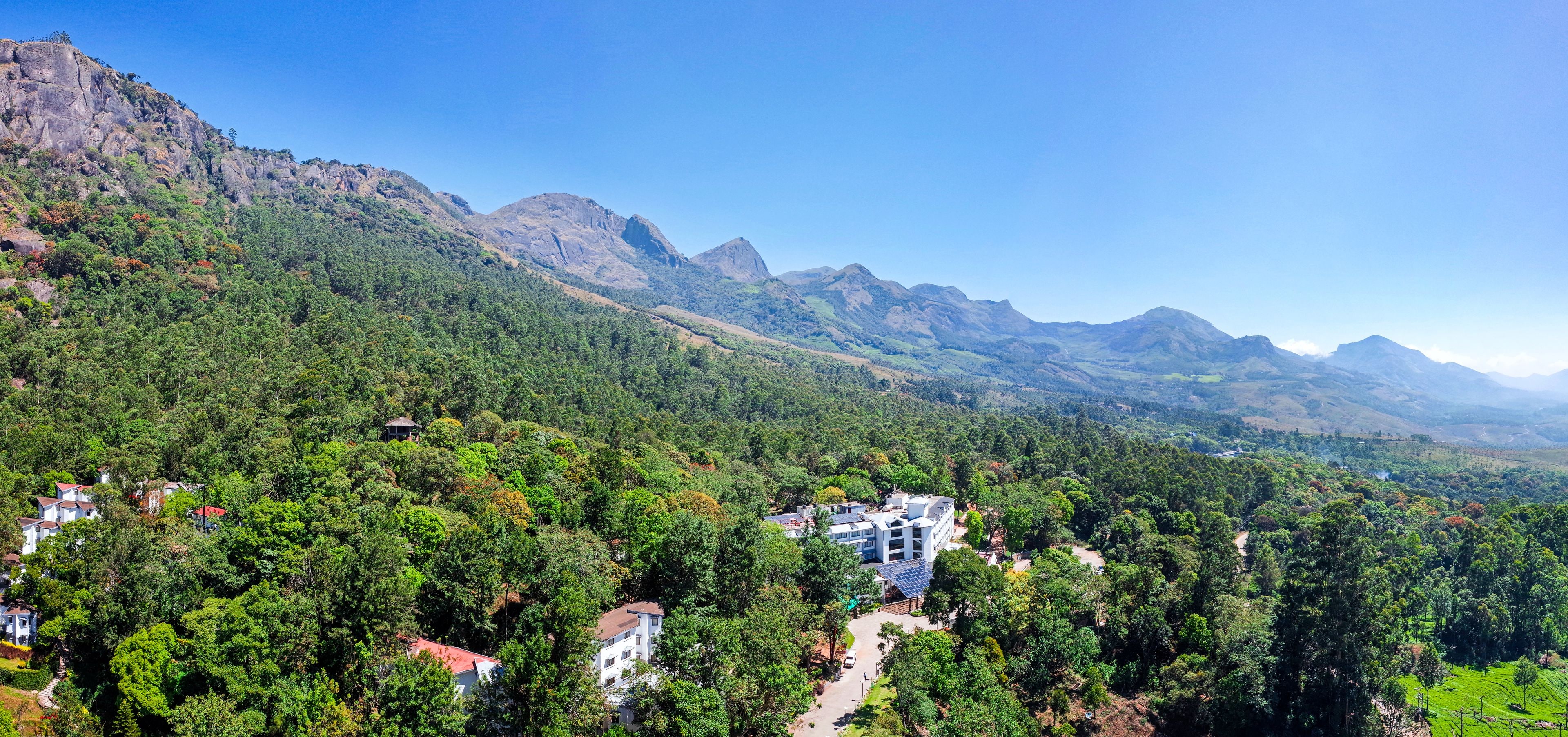 A mountainous landscape with a building featuring multiple floors and solar panels, surrounded by dense forest and residential houses.