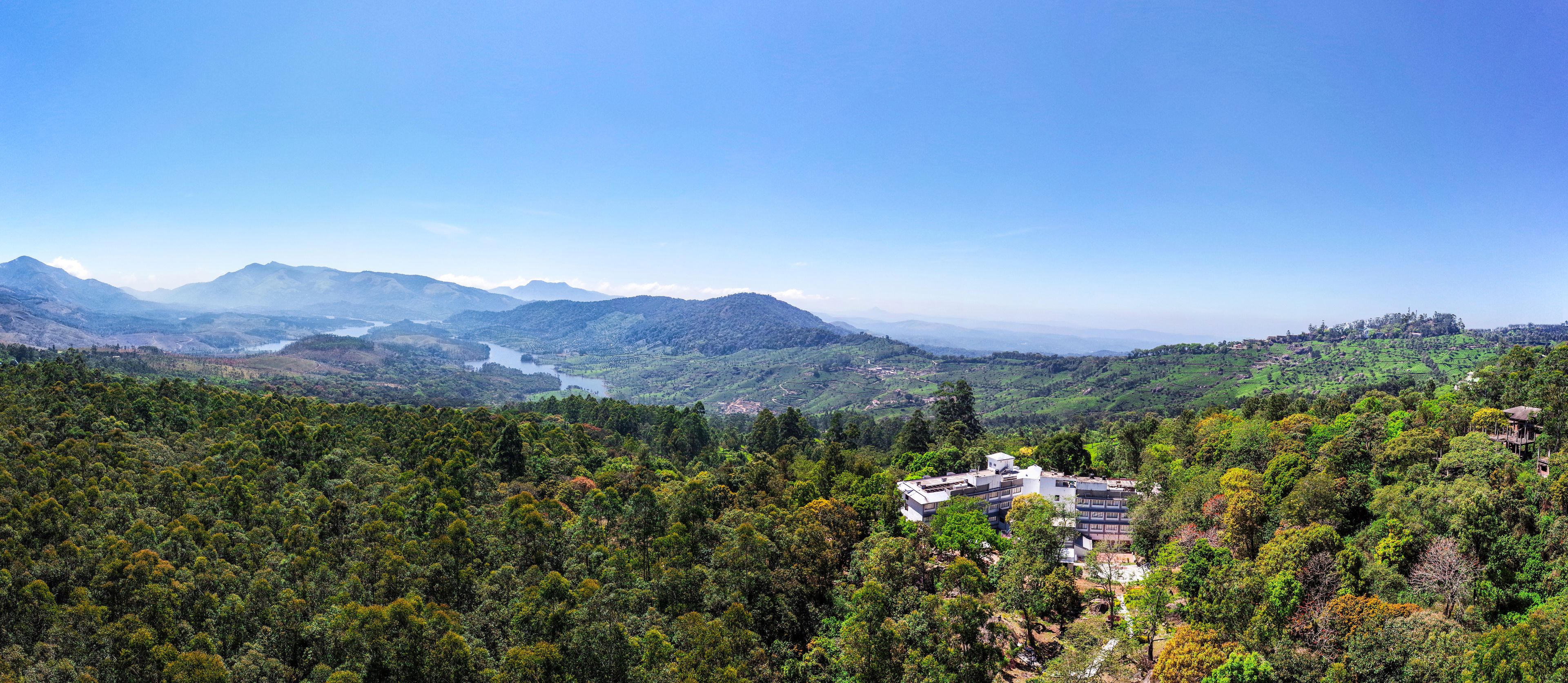 A mountainous landscape with a modern building surrounded by dense forest, overlooking a valley and distant mountains.