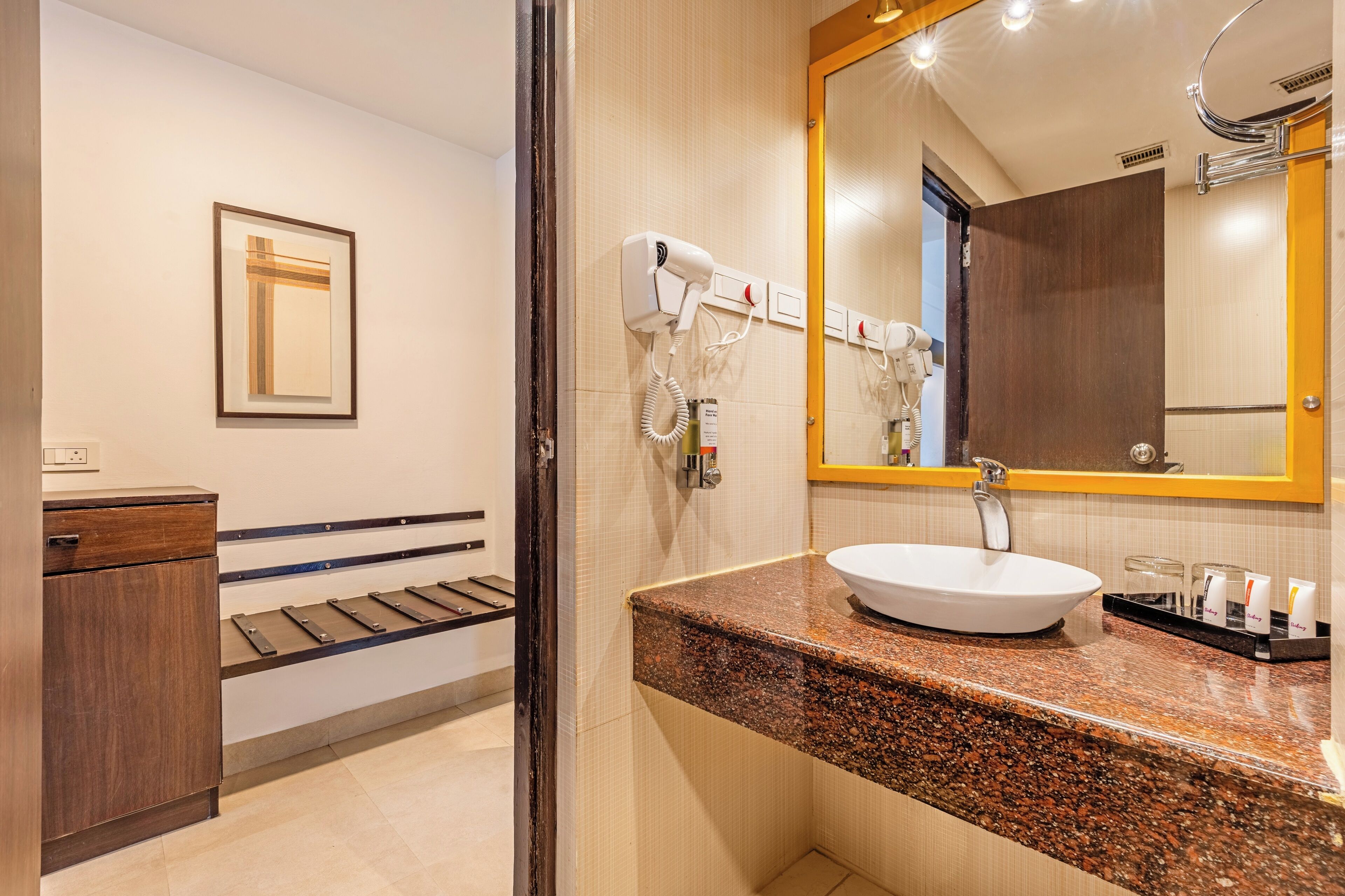 A modern bathroom with a granite countertop, a white sink, and a large mirror. There are hair dryers mounted on the wall and a wooden bench with a towel rack.