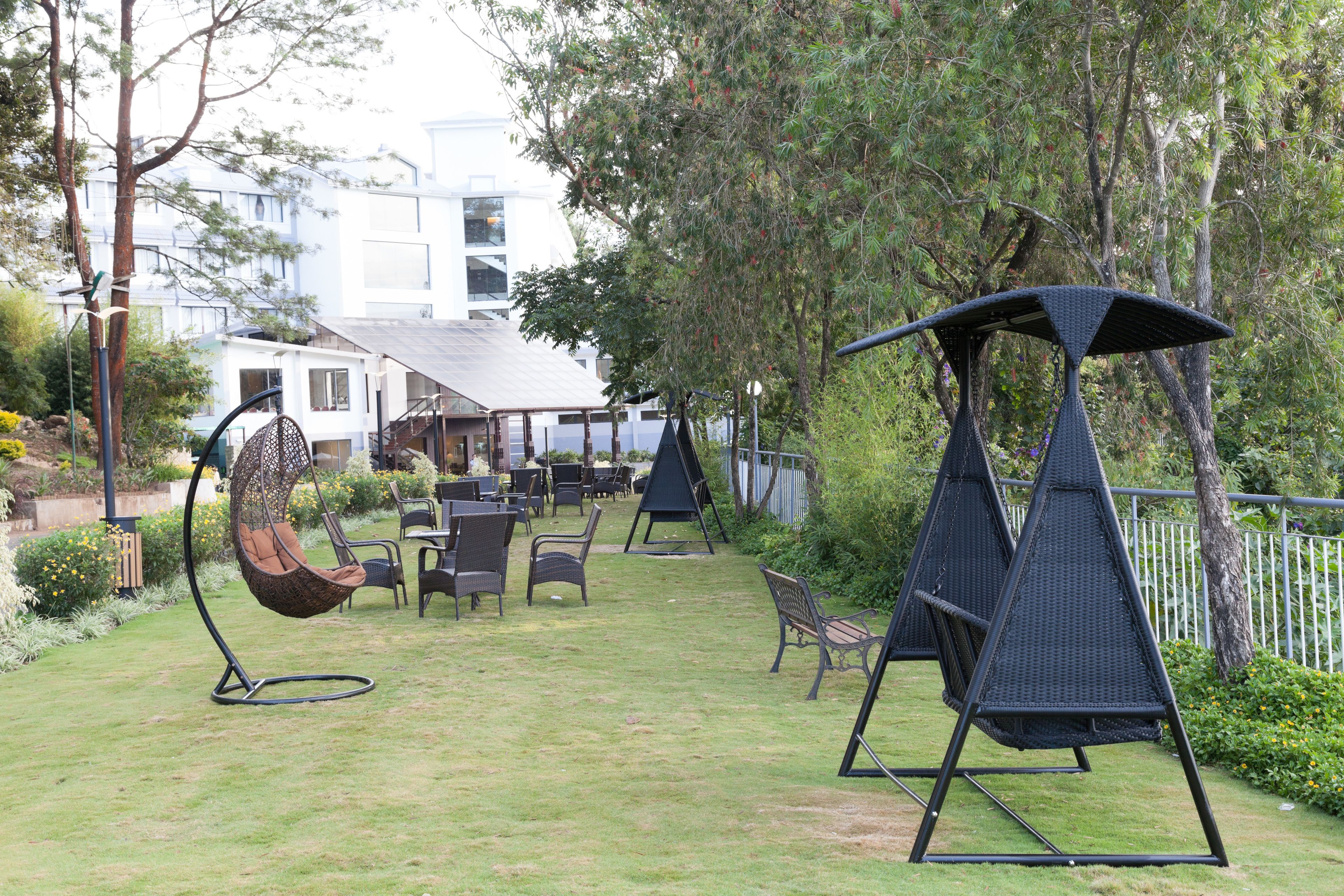 A patio area with outdoor seating, including a swing chair and a swing set, surrounded by greenery and a building in the background.