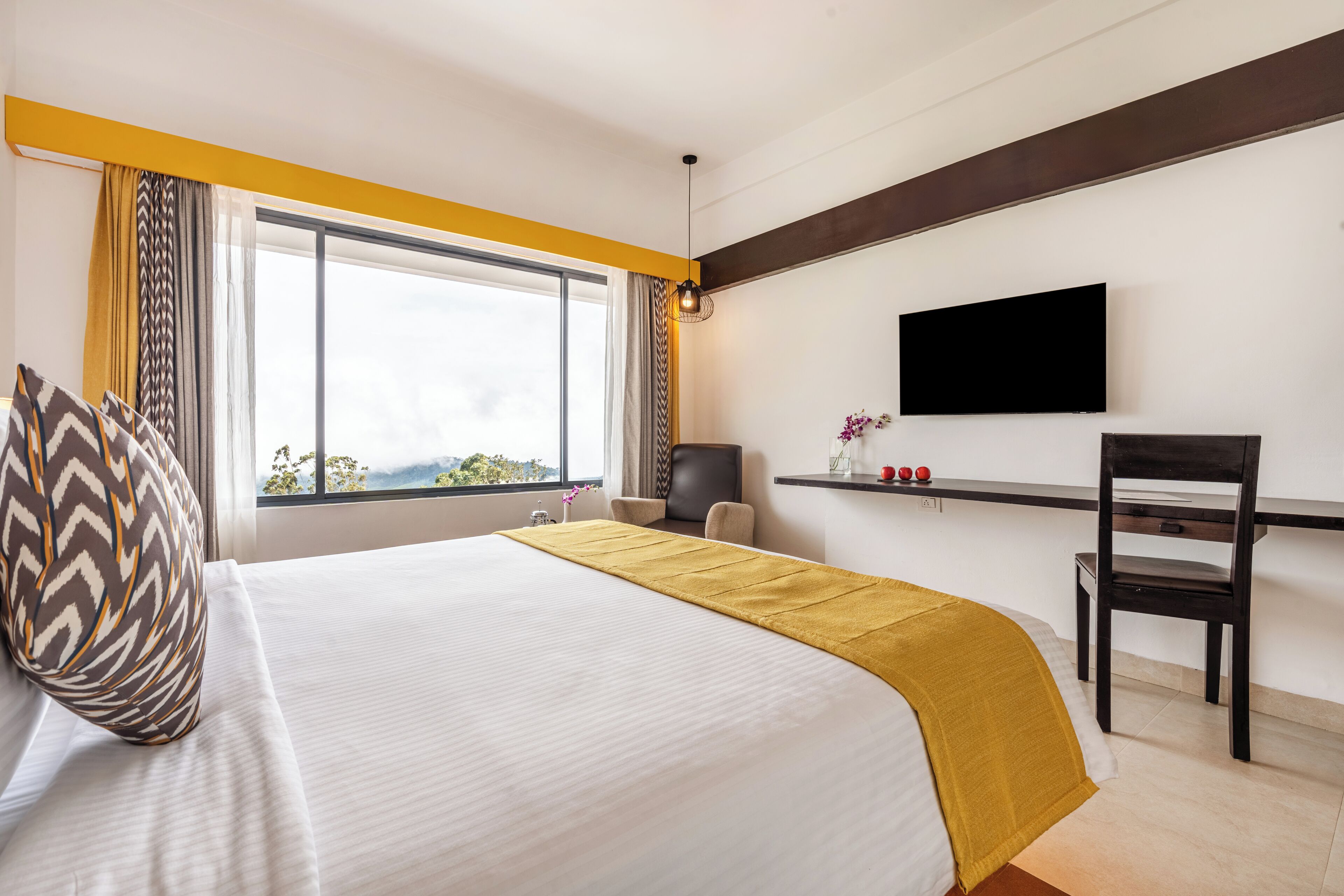 A hotel room with a large bed, a desk with a chair, a TV mounted on the wall, and a window with curtains.