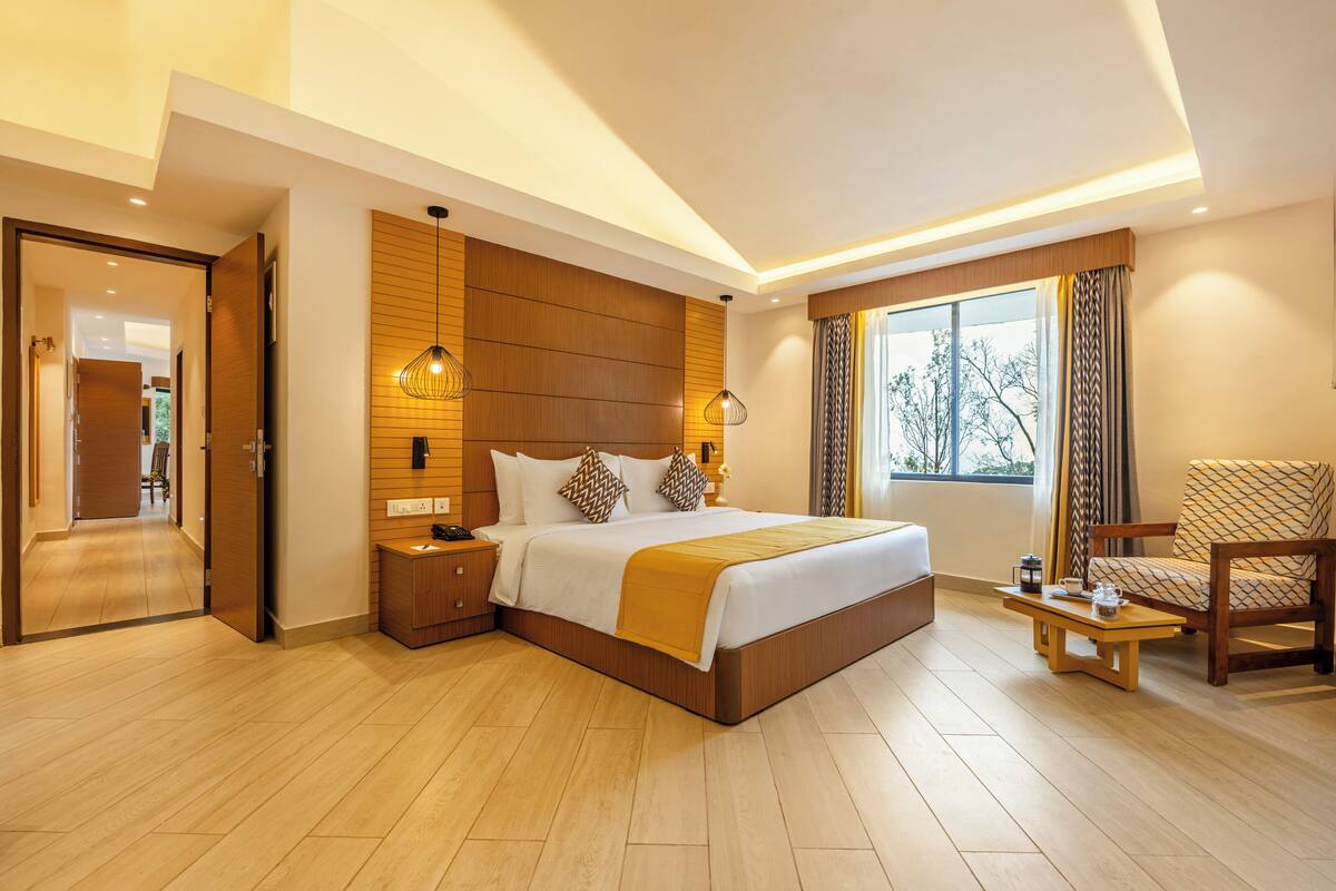A modern hotel room with a large bed, wooden paneling, a sitting area with a chair, and a view of the outdoors through the window.