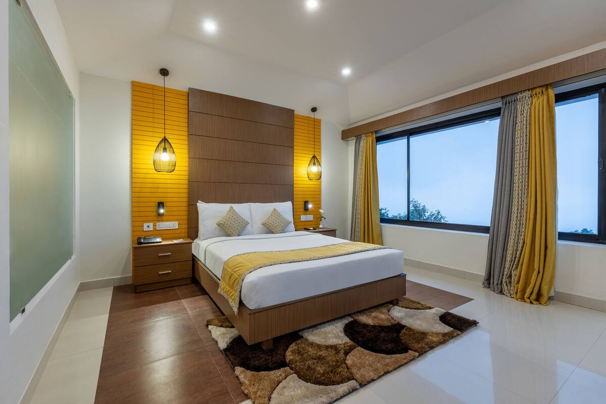 A modern bedroom with a large bed, wooden panel wall, two bedside lamps, and a view of the sky through large windows.