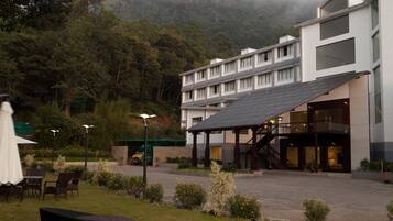 A hotel with a modern design, featuring a covered outdoor seating area, a staircase leading to the upper levels, and a view of a mountain in the background.