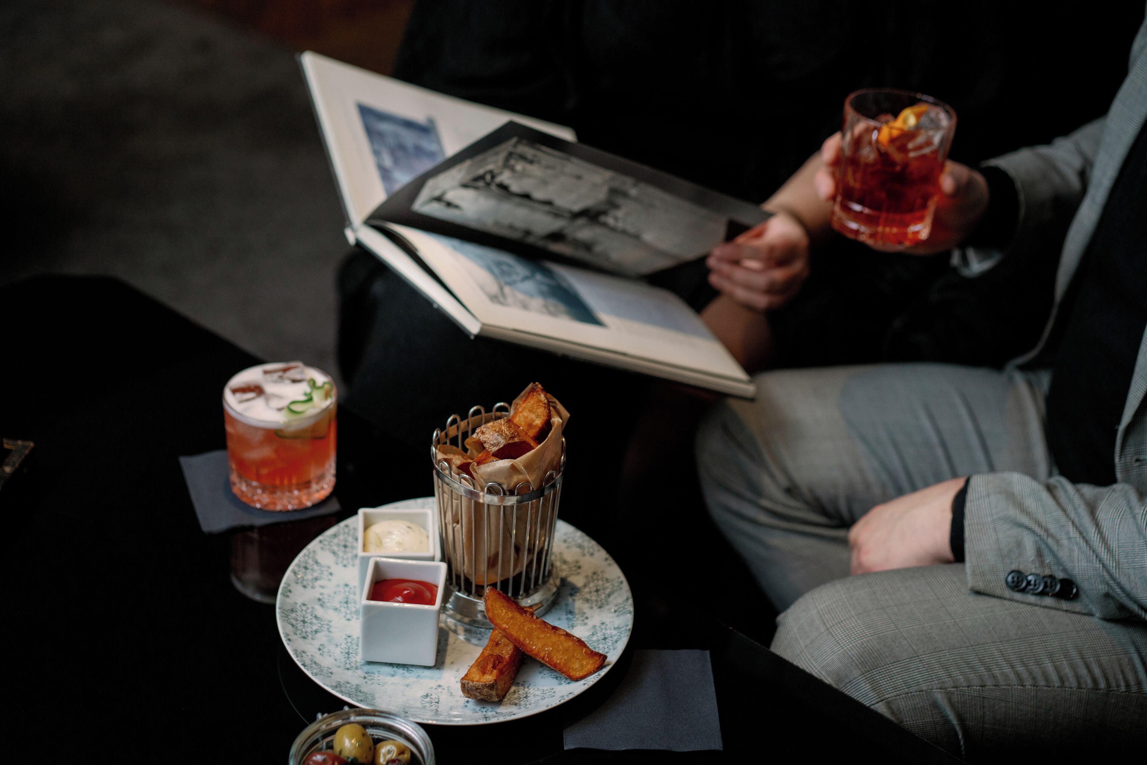 A person holding a drink and a book, with a plate of food and a glass of beverage on a table.