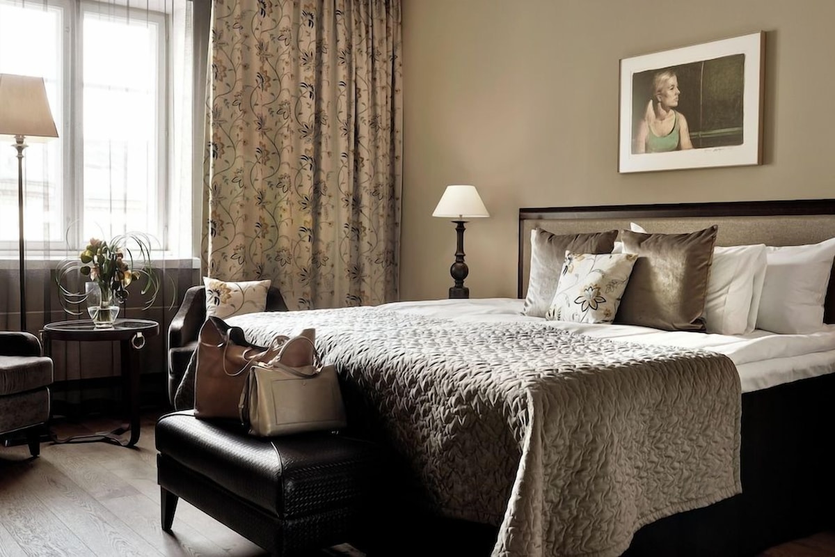 A hotel room with a bed, a nightstand, a lamp, a chair, a bench, a window with curtains, and a framed picture on the wall.