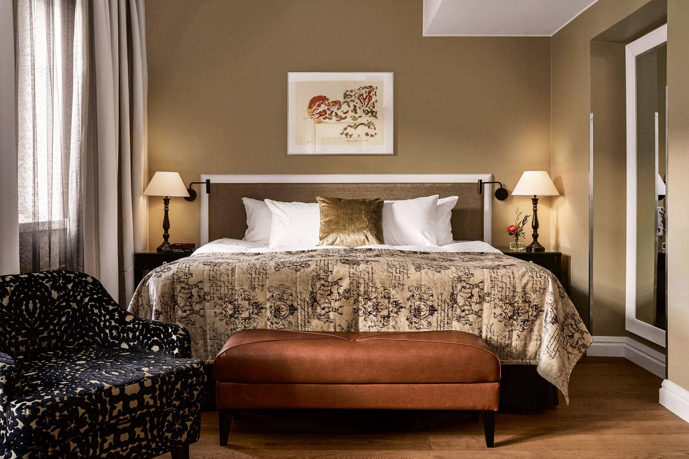 Habitación de hotel con cama, dos lámparas de noche, un cuadro en la pared, colcha estampada, un banco marrón y una silla con diseño de estrellas.