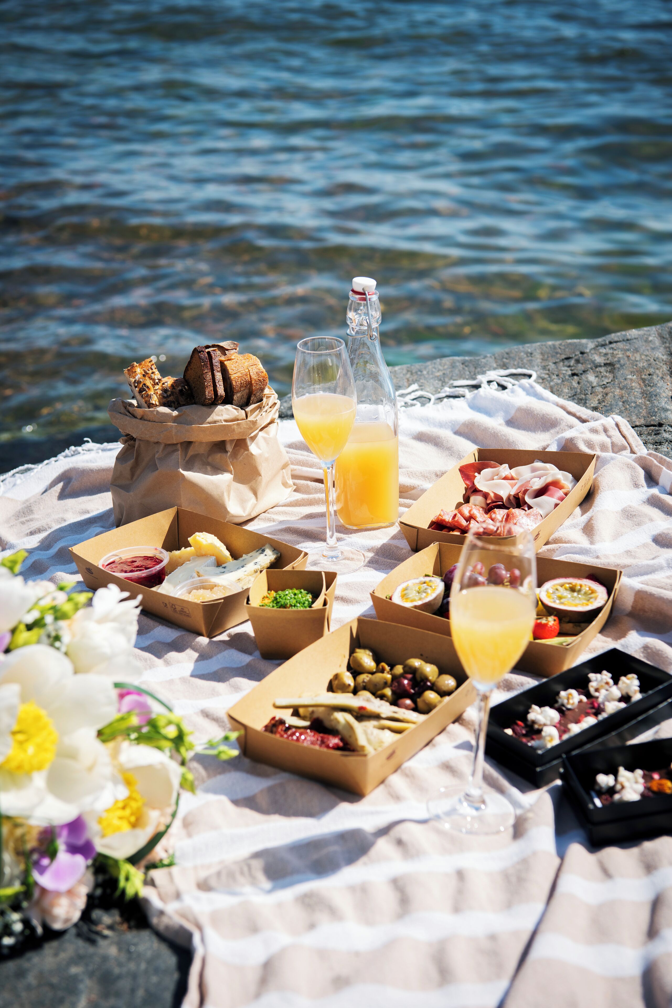 A picnic setup with various food items and drinks on a blanket near a body of water.