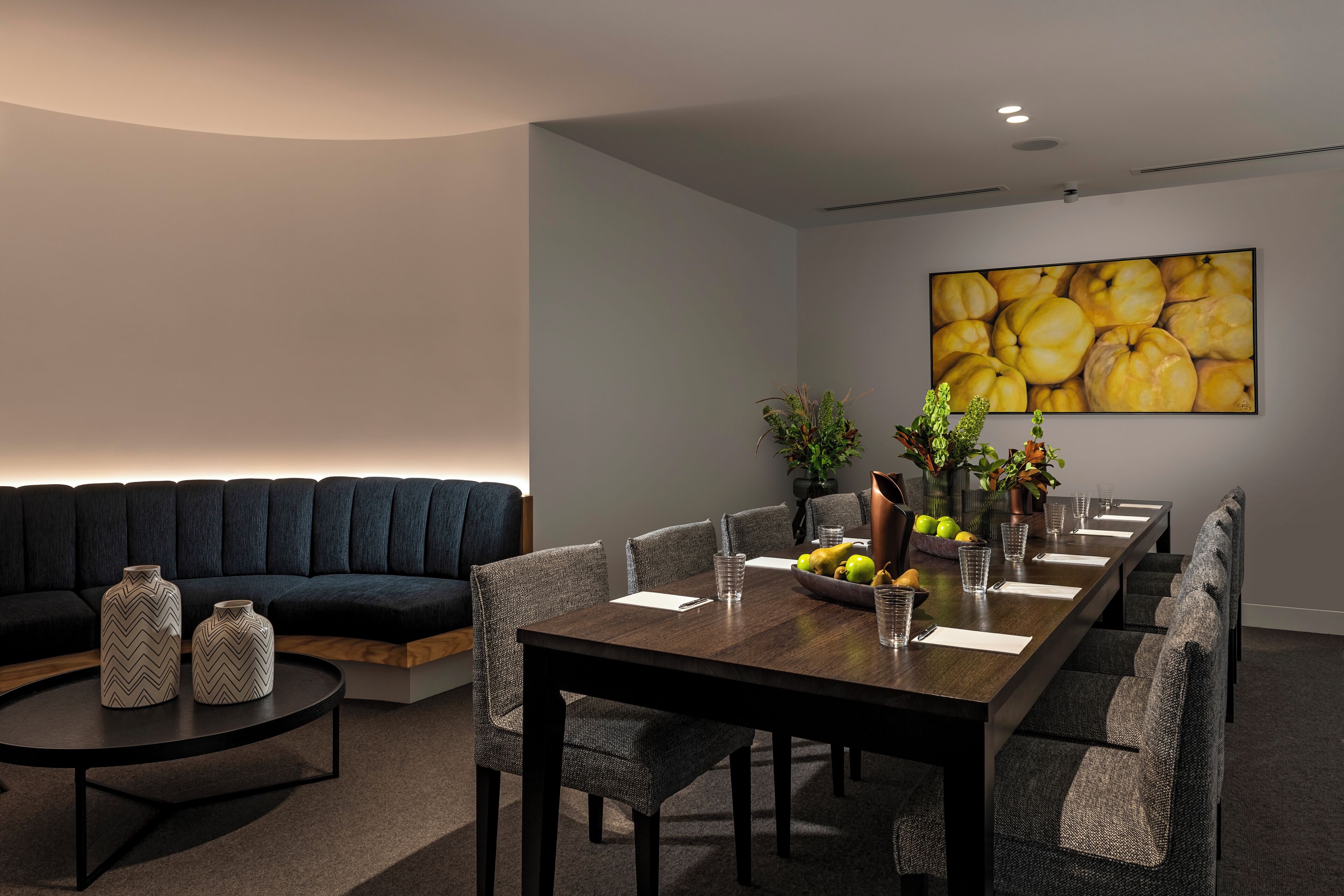 A modern dining area with a dark sofa, a round coffee table, and a long dining table set with chairs, vases, and a fruit bowl.