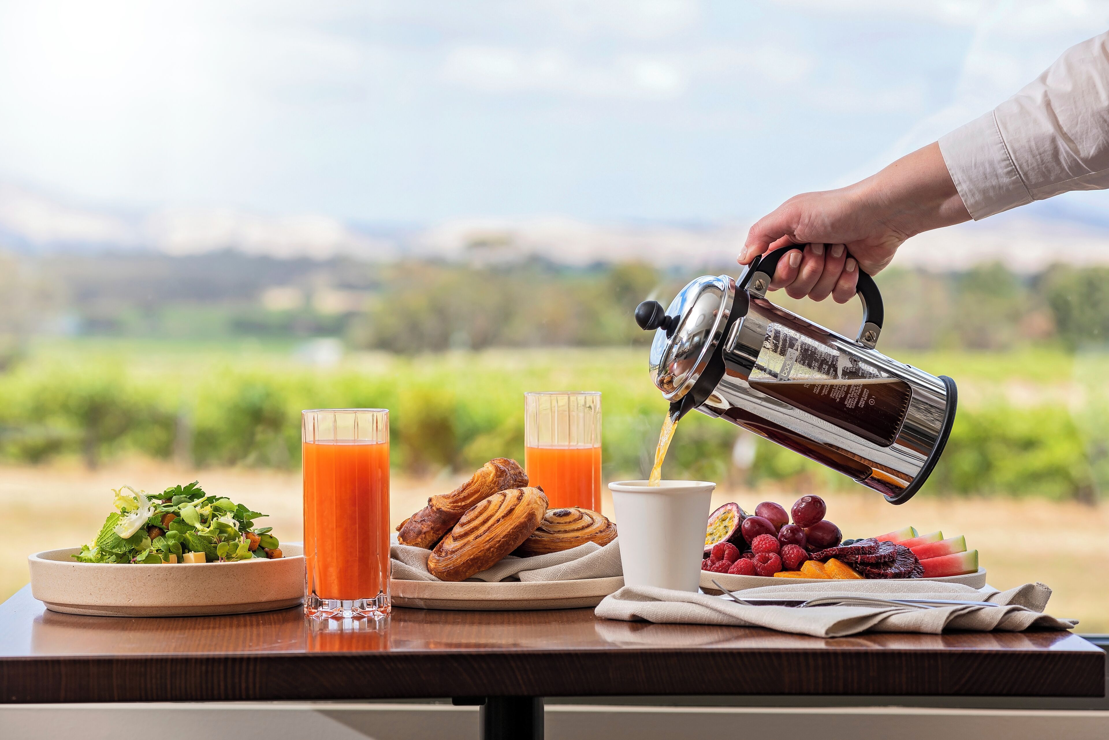 Ein Frühstückstisch mit einer Kaffeekanne, die Kaffee in eine Tasse gießt, zwei Orangensaftgläsern, Gebäck und einer Schüssel mit Salat.