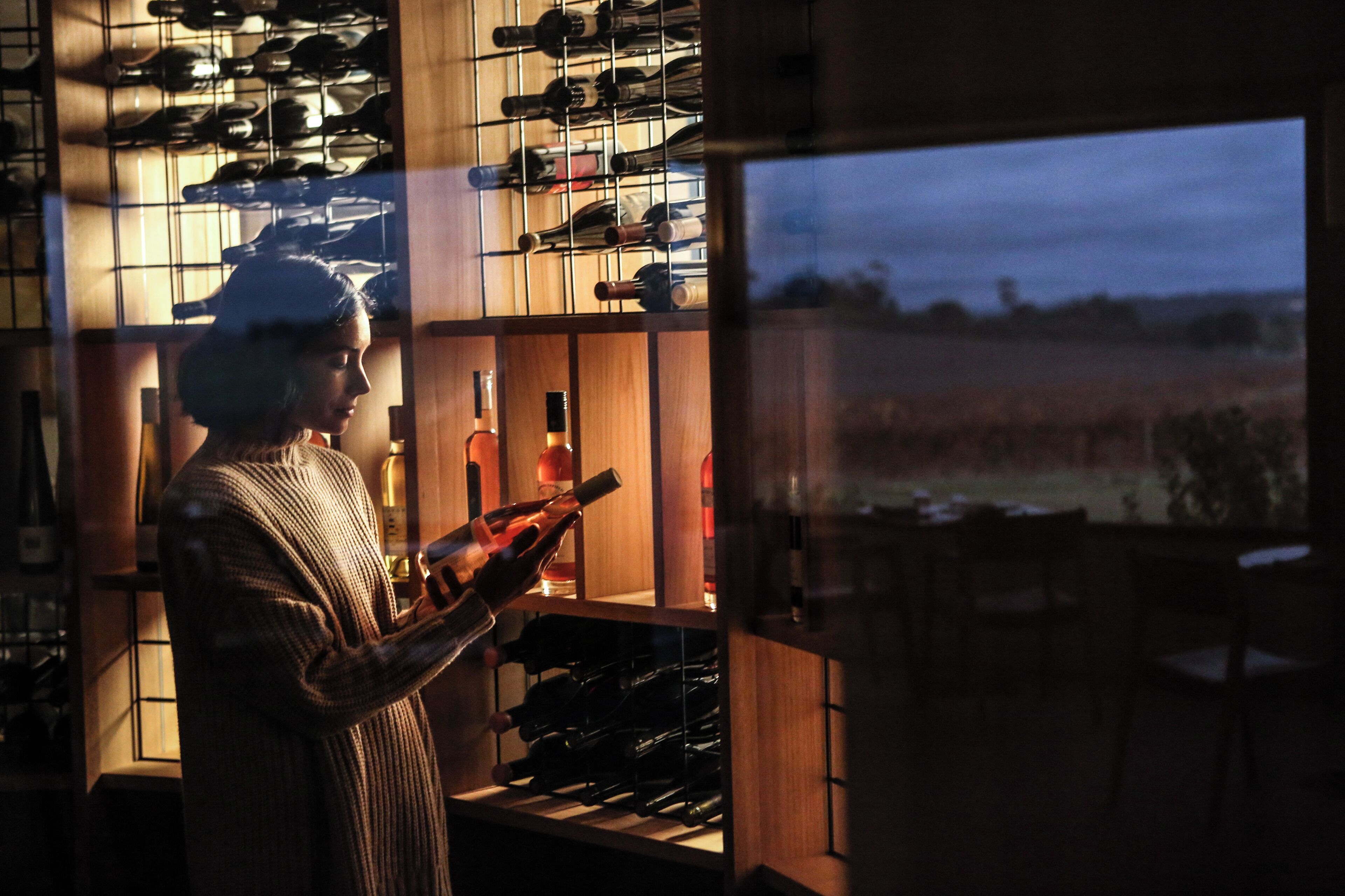 A person holding a wine bottle in a dimly lit room with wooden shelves filled with wine bottles.