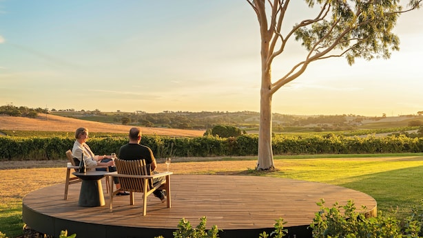 Una terraza de madera circular con dos personas sentadas en una mesa, rodeada de follaje y vistas a un paisaje montañoso.