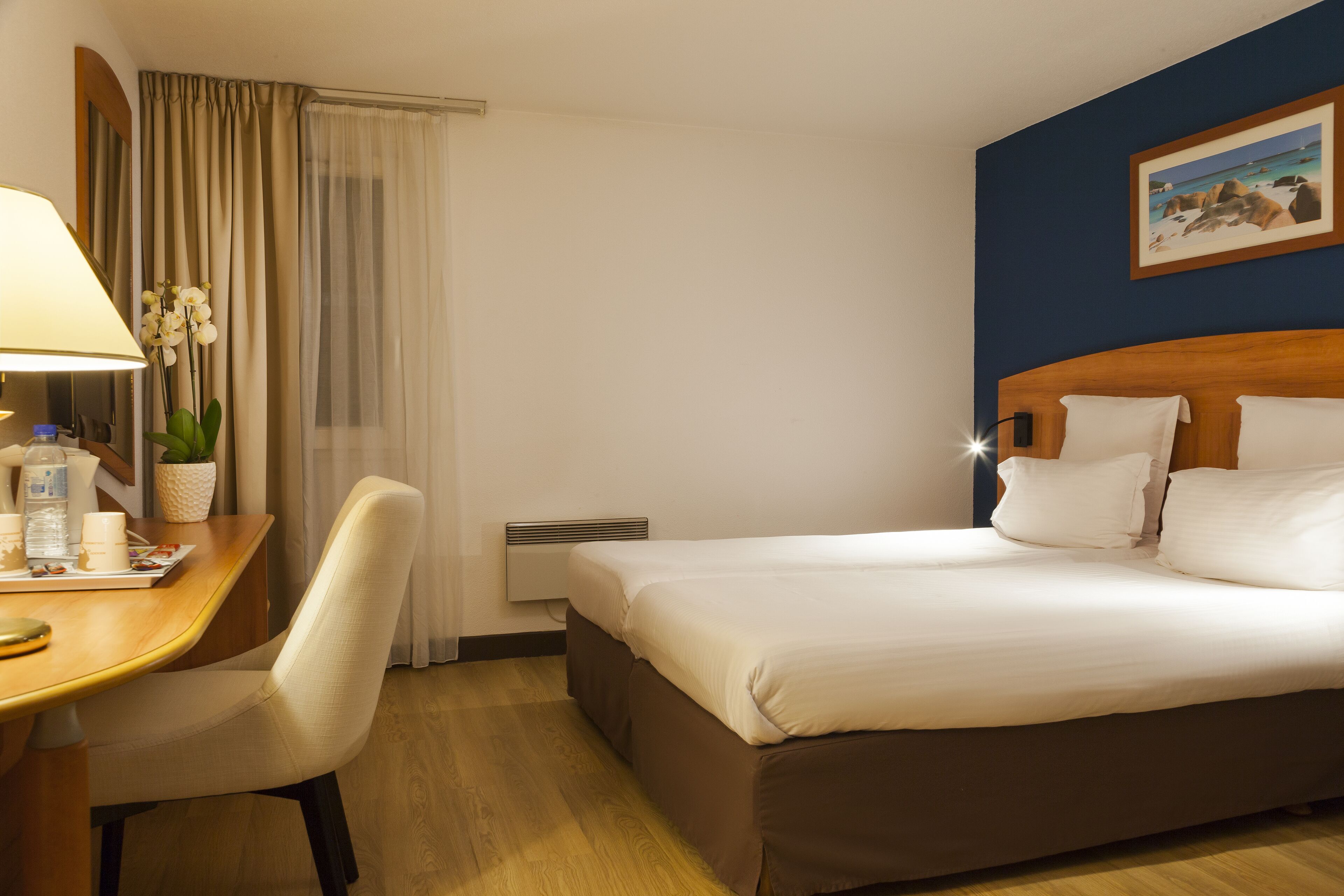 A hotel room with a bed, a desk, a chair, a lamp, a potted plant, and a painting on the wall.