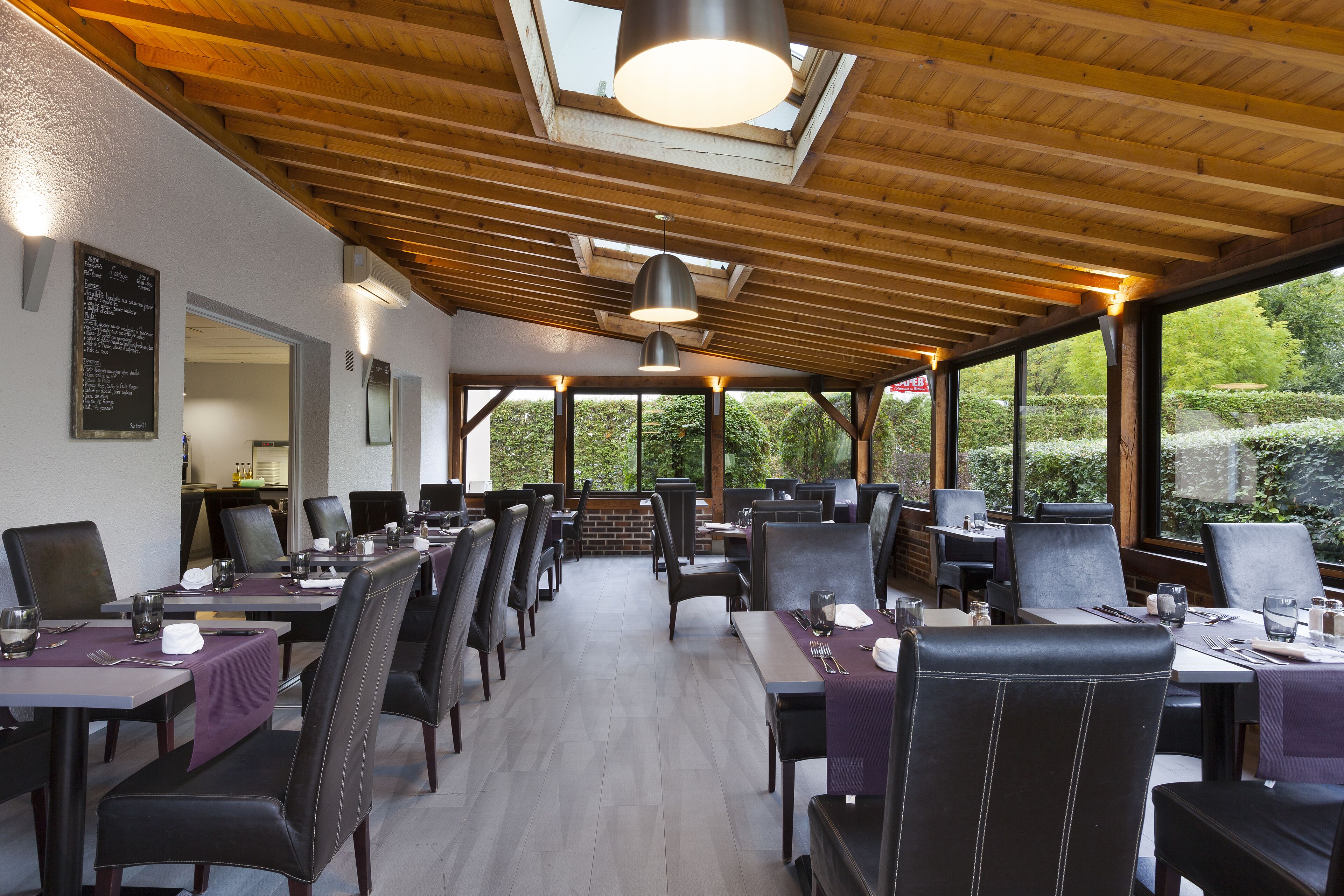 Ein Restaurant mit einer langen Essecke, Holzbalkendecke und großen Fenstern mit Blick auf Grünflächen.