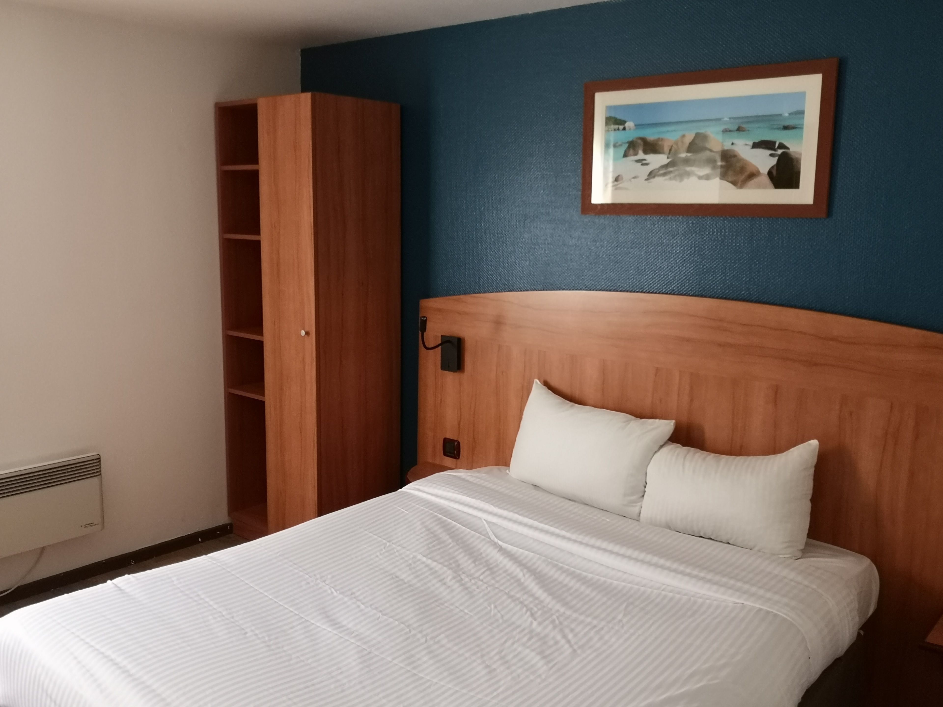 Una camera d'albergo con una testiera in legno, una coperta bianca, un cuscino bianco, un armadio in legno e un quadro appeso al muro.