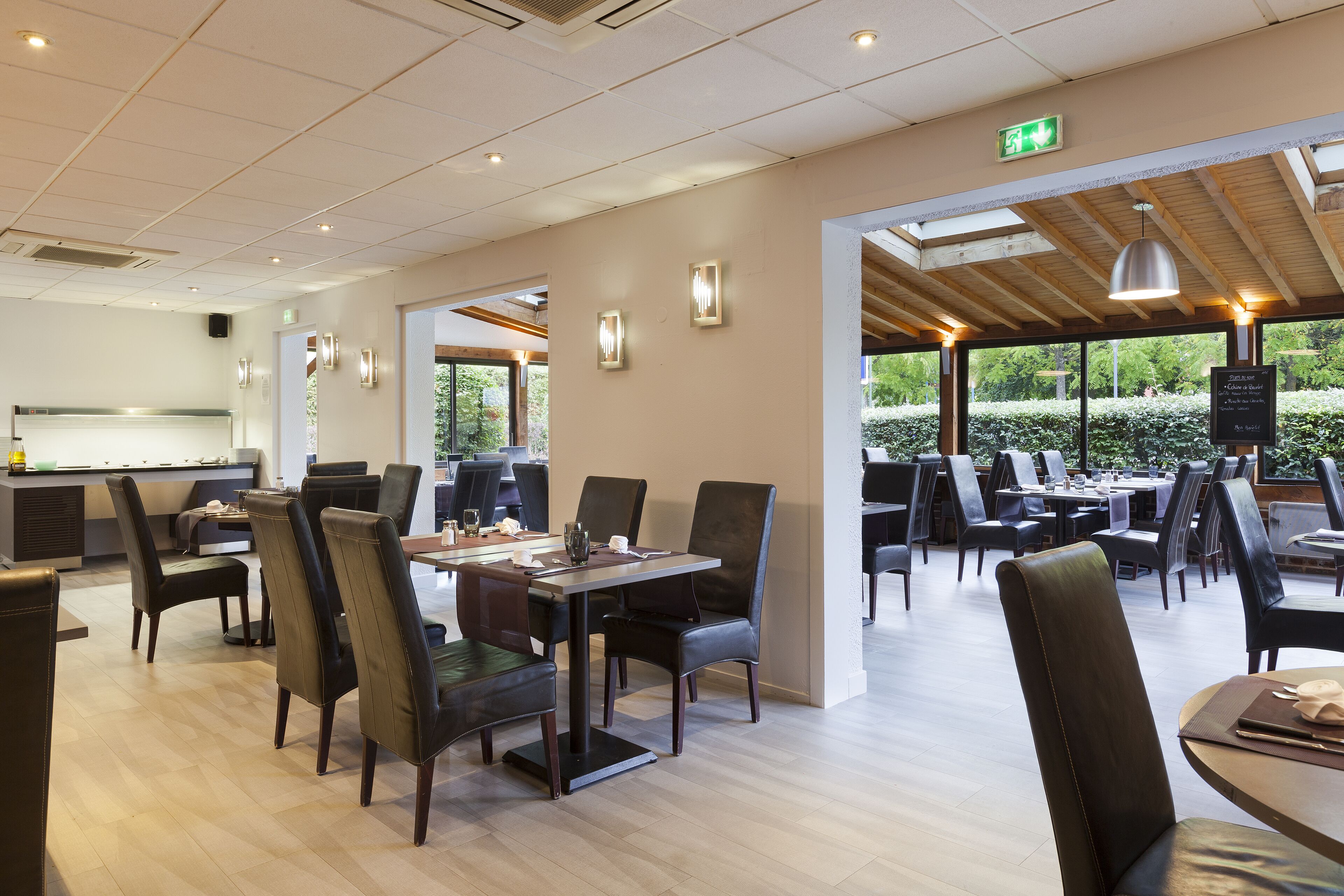 Een modern restaurant met meerdere eetgedeeltes, houten balken in het plafond en grote ramen met uitzicht op groen.