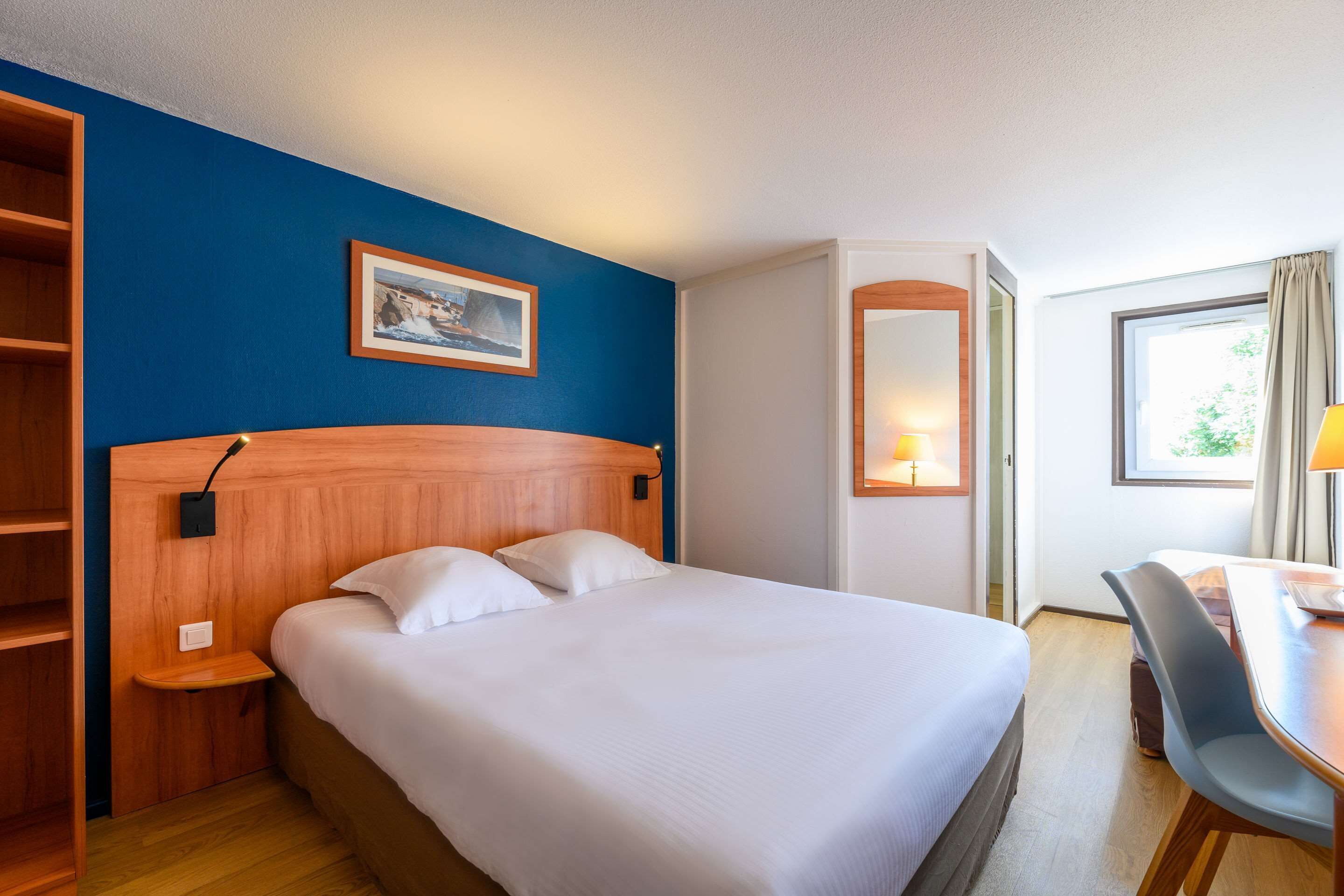 A hotel room with a wooden headboard, a bed with white linens, a desk with a chair, a window with curtains, and a framed picture on the wall.