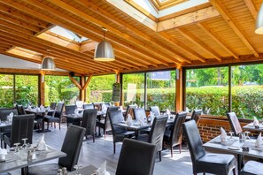 Un área de comedor al aire libre con vigas de madera, mesas dispuestas con vajilla blanca y vistas a la vegetación.