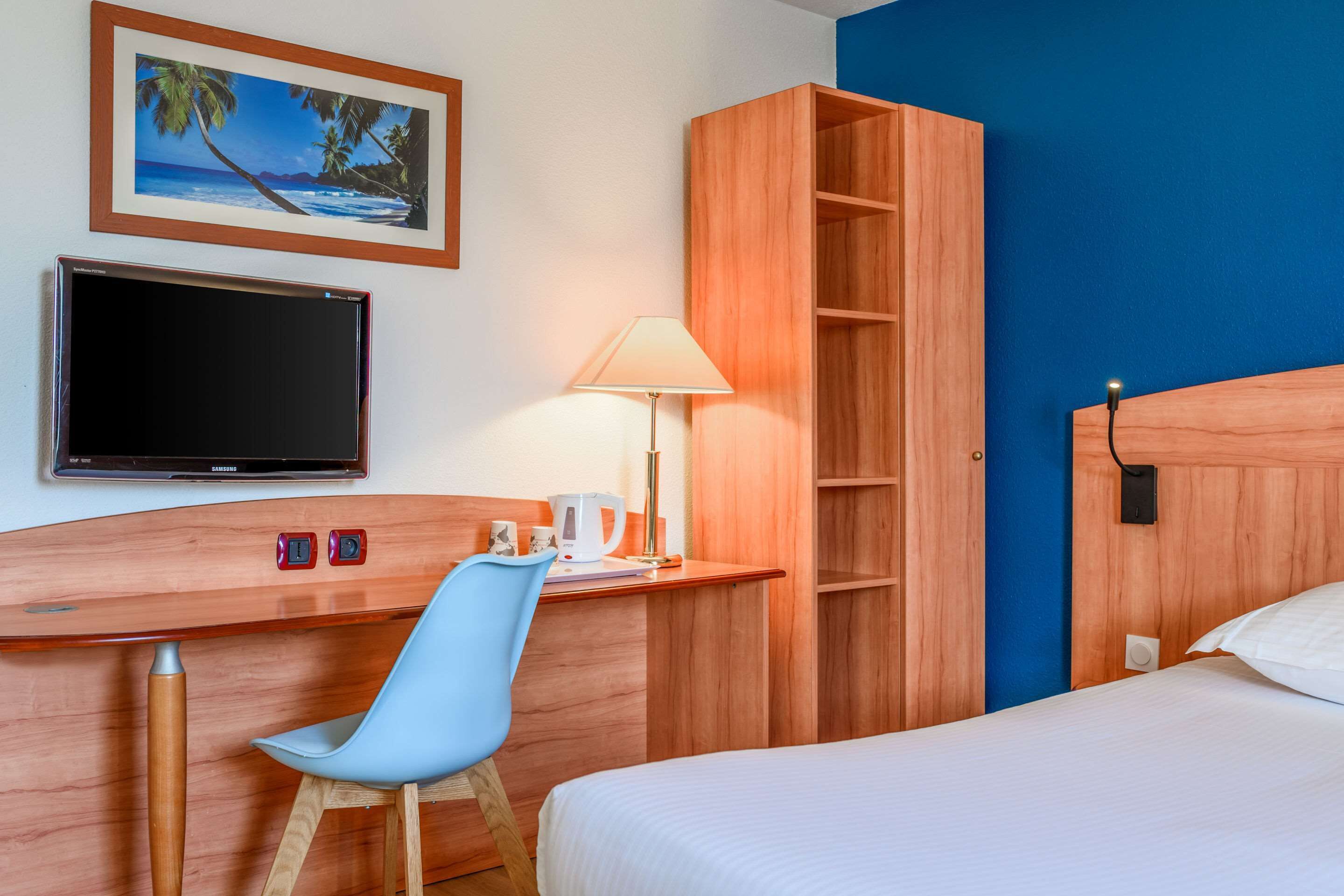 Een hotelkamer met een bed, een bureau met stoel, een televisie, een lamp en een houten garderobe.