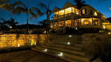 A large, two-story house with a stone wall entrance, multiple balconies, and a well-lit outdoor area.