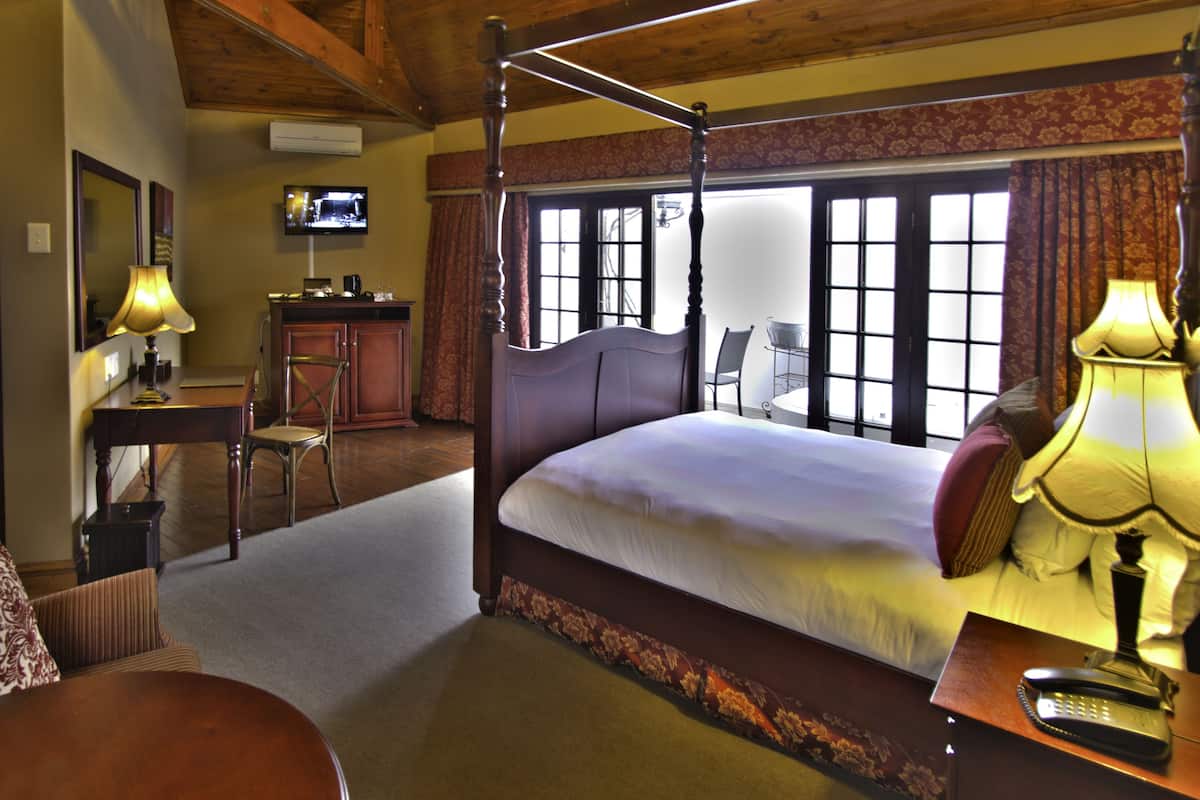 A four-poster bed with a television above it, a desk with a lamp, and a chair in a room with large windows.