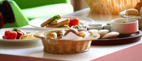 A breakfast setting with various food items on a table, including bread, pastries, and a plate with tomatoes and herbs.