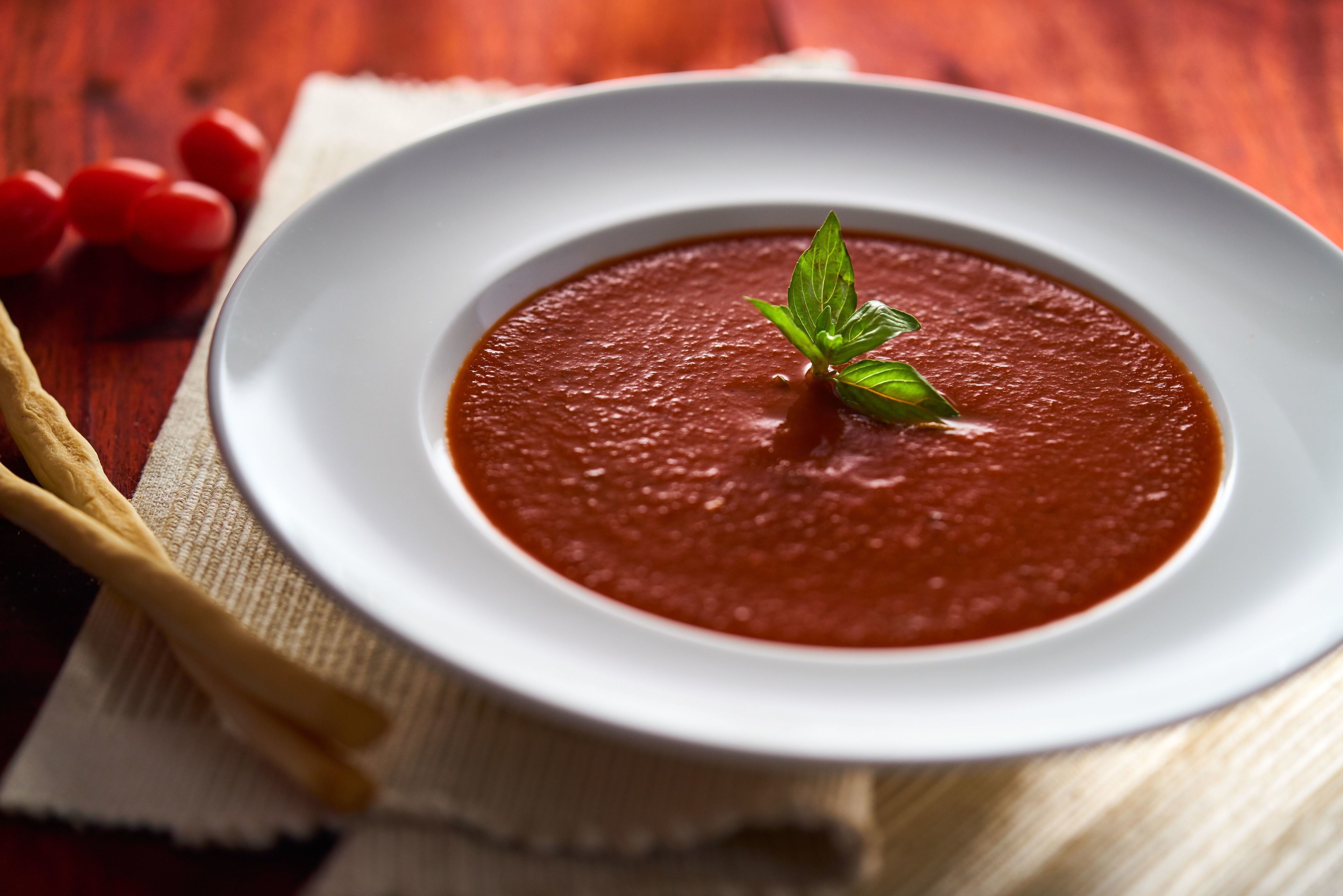 A bowl of tomato soup on a white plate with a basil garnish, placed on a wooden table with a white napkin and a wooden spoon.