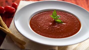 A bowl of tomato soup on a white plate with a basil garnish, placed on a wooden table with a white napkin and a wooden spoon.