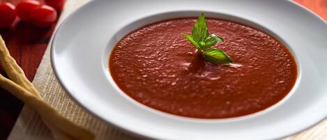 A bowl of tomato soup on a white plate with a basil garnish, placed on a wooden table with a white napkin and a wooden spoon.