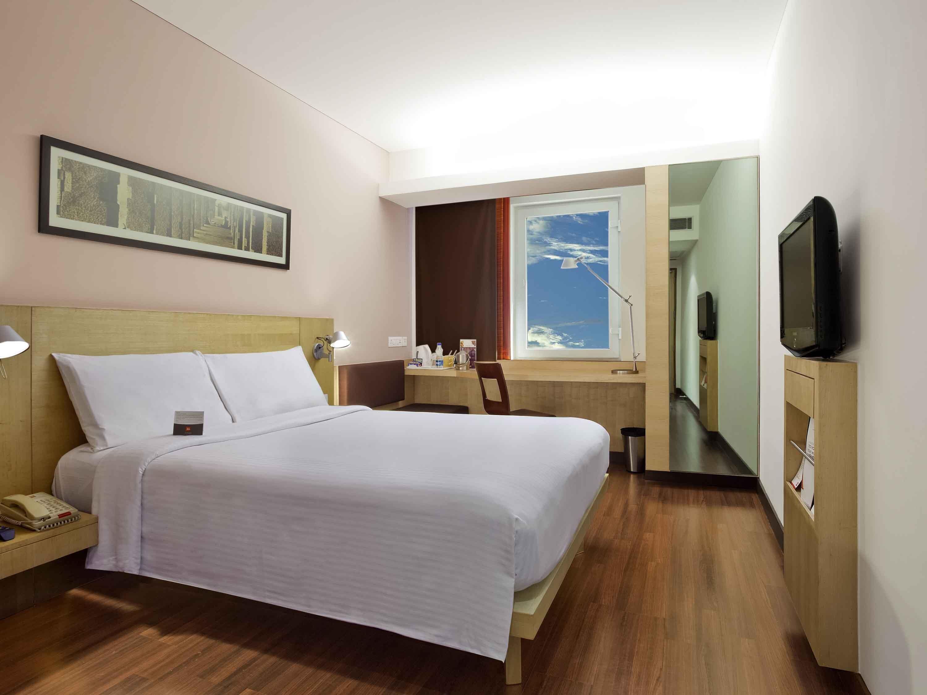 A hotel room with a bed, bedside table with a lamp, a desk with a chair, a TV, and a window with a view of the sky.