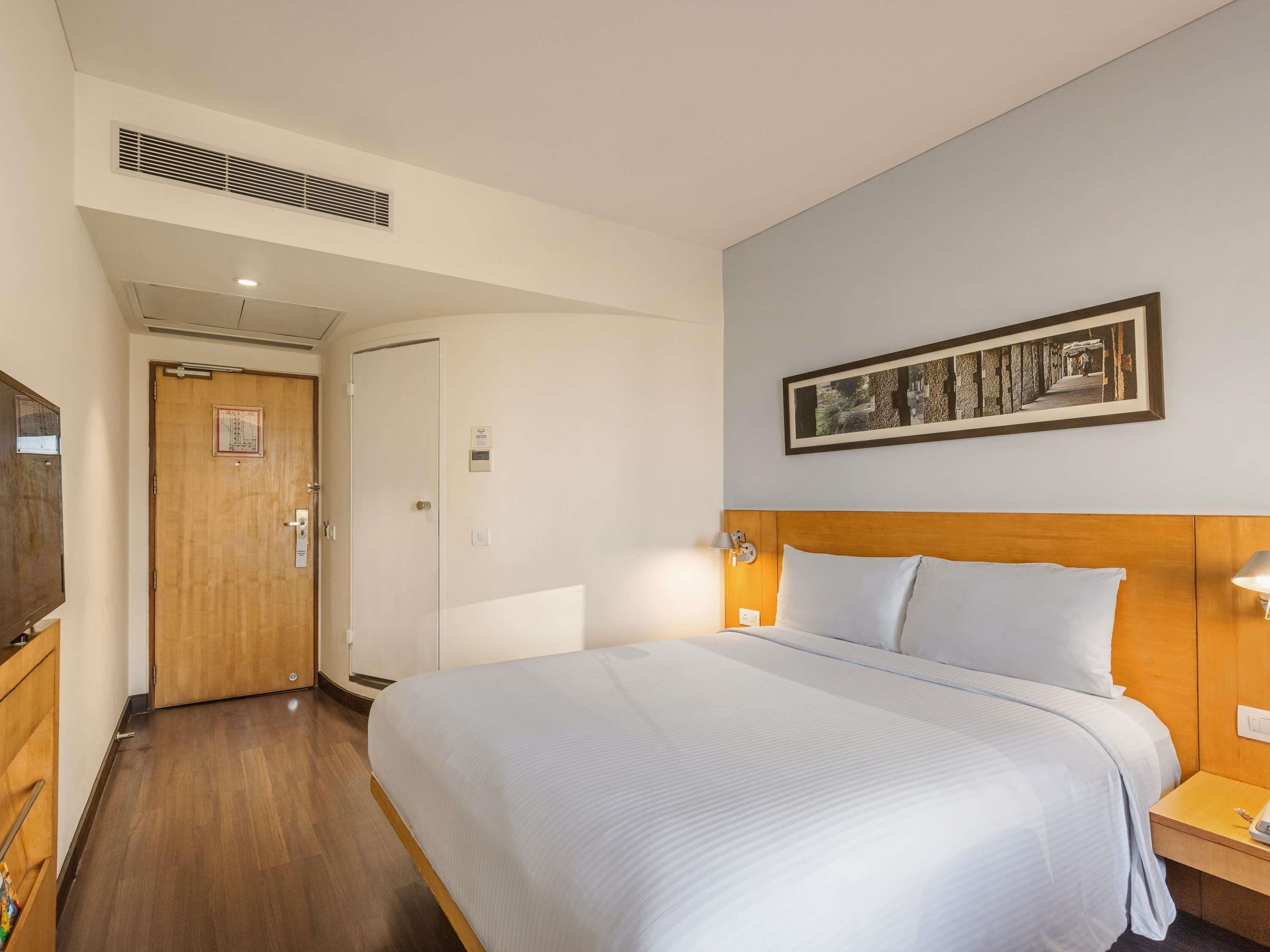 A hotel room with a bed, bedside lamps, a wooden headboard, a framed picture on the wall, a door, and a vent on the ceiling.