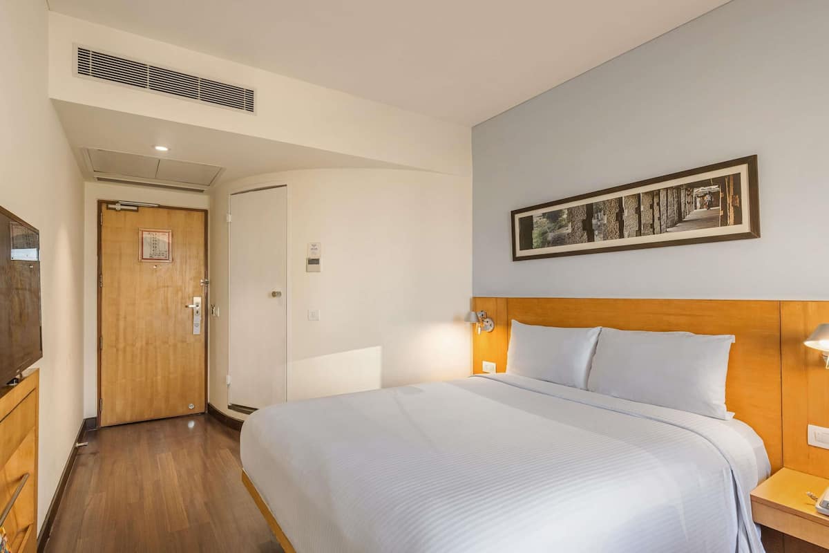 A hotel room with a bed, bedside lamps, a wooden headboard, a framed picture on the wall, a door, and a vent on the ceiling.