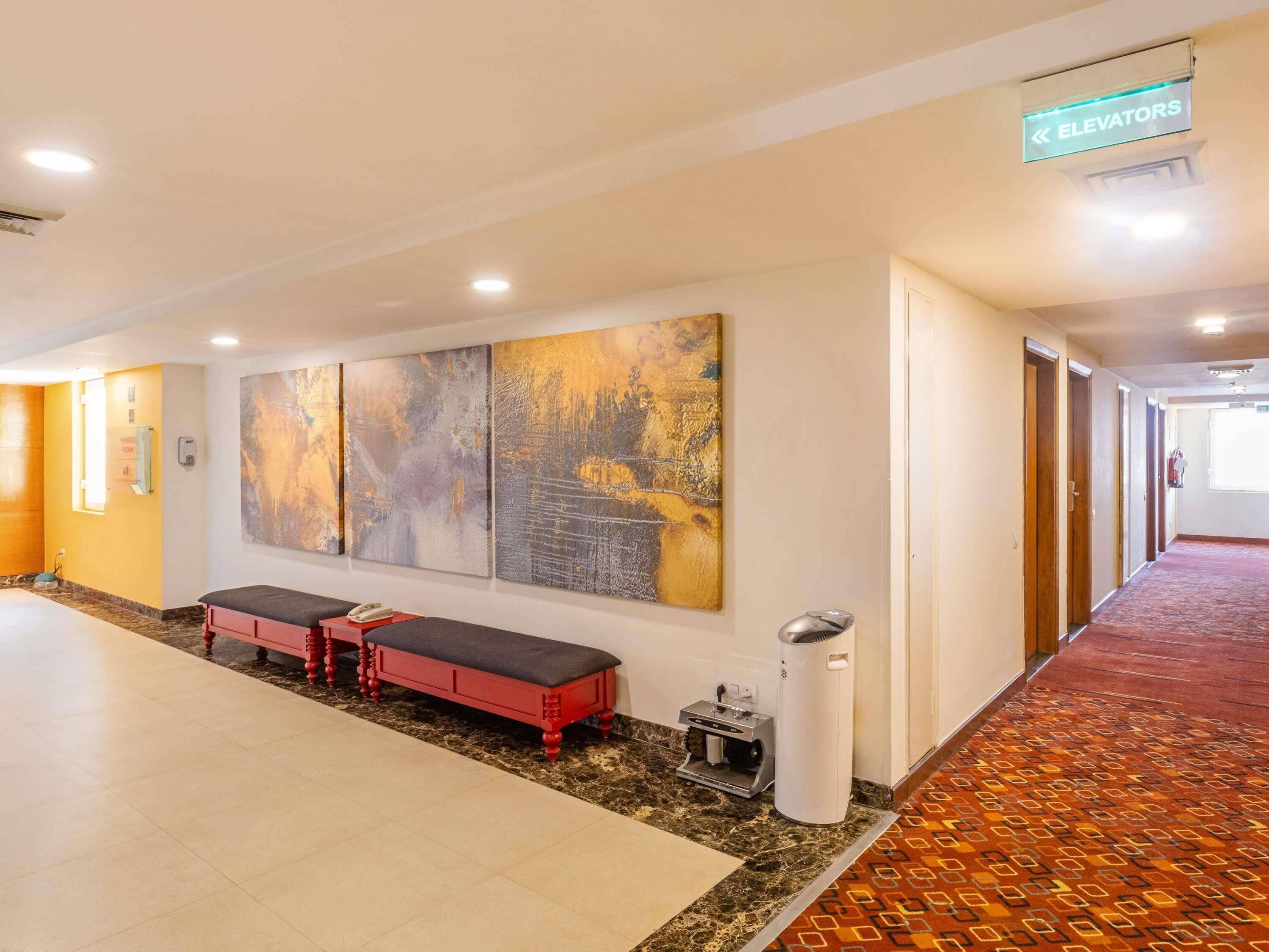 A hallway with a bench, artwork, an elevator sign, and a carpeted floor.
