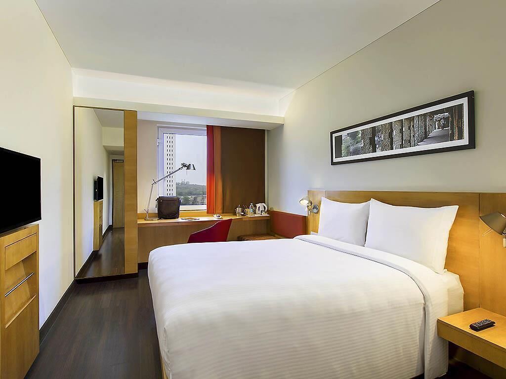 A hotel room with a large bed, a desk, a chair, a TV, a framed picture on the wall, and a window with curtains.