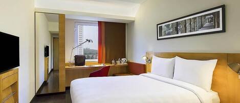 A hotel room with a large bed, a desk, a chair, a TV, a framed picture on the wall, and a window with curtains.