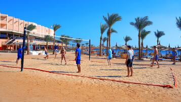 A beach volleyball game on a sandy court with palm trees and a building in the background.