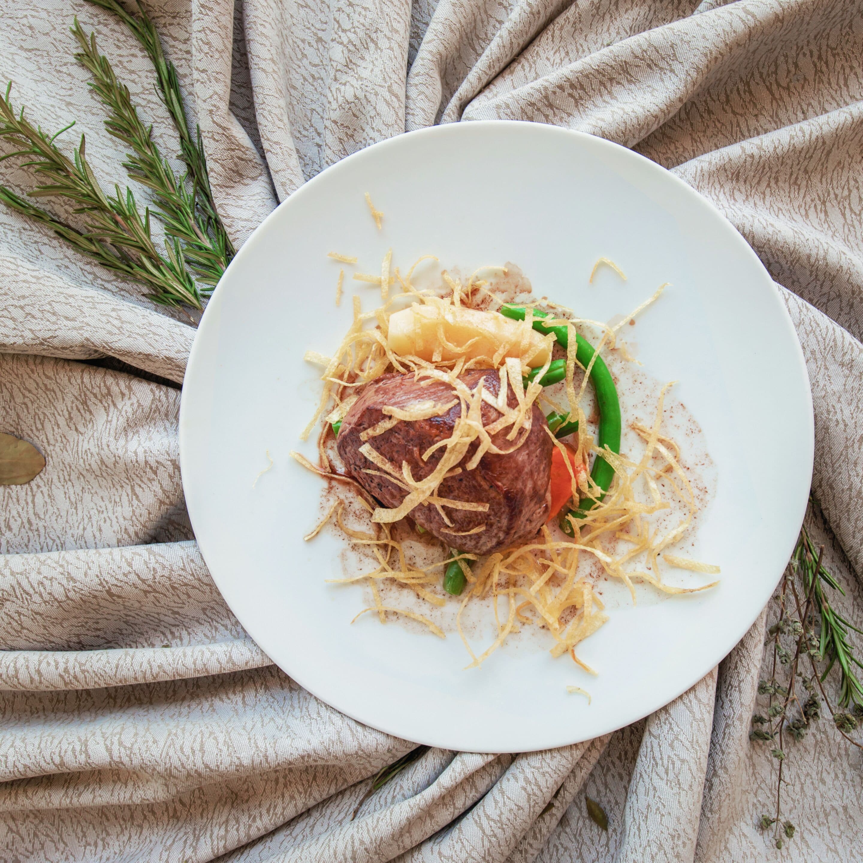 A white plate with a meat dish, garnished with green vegetables and shredded cheese, on a textured beige cloth.